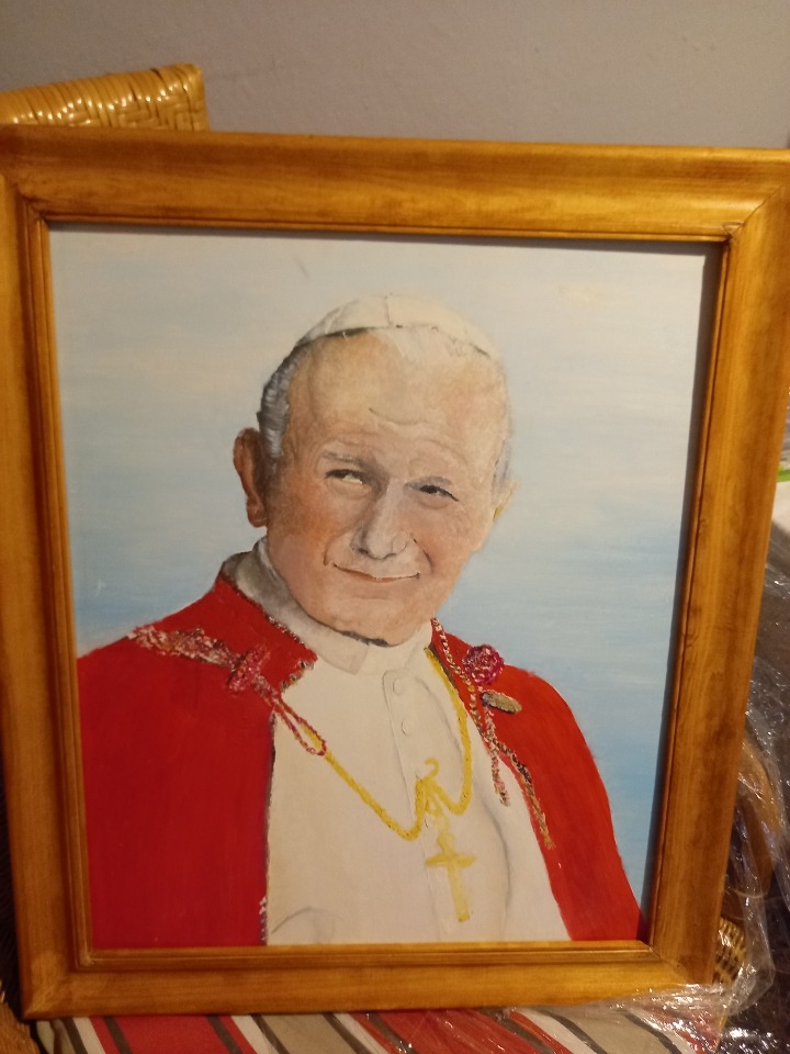Portret Jana Pawła II w złotej ramie, malowany farbami olejnymi, z widocznymi pociągnięciami pędzla, ustawiony na wzorzystym materiale.