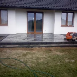 Nowo ułożony taras z szarej kostki brukowej przed domem z białymi ścianami i brązowymi oknami, z ubijarką wibracyjną obok, w tle pochmurne niebo.