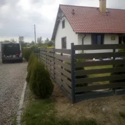 Nowoczesne ogrodzenie panelowe z kompozytu w kolorze ciemnego drewna, zamontowane na posesji z trawnikiem i młodymi tujami, na tle domu z czerwoną dachówką i zaparkowanego busa.