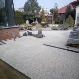 Ułożona jasna nawierzchnia z kostki brukowej z widocznym odpływem liniowym, przygotowane stosy kostki do dalszych prac, widok na ogród i dom w tle.