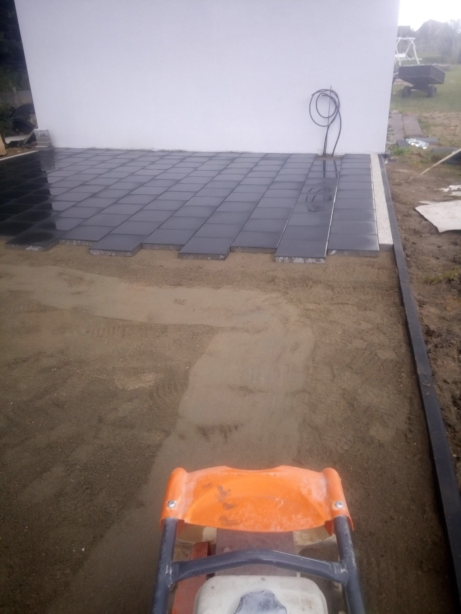 Proces zagęszczania podłoża pod czarne płyty chodnikowe przy białej ścianie budynku, widoczny zagęszczarką wibracyjną.