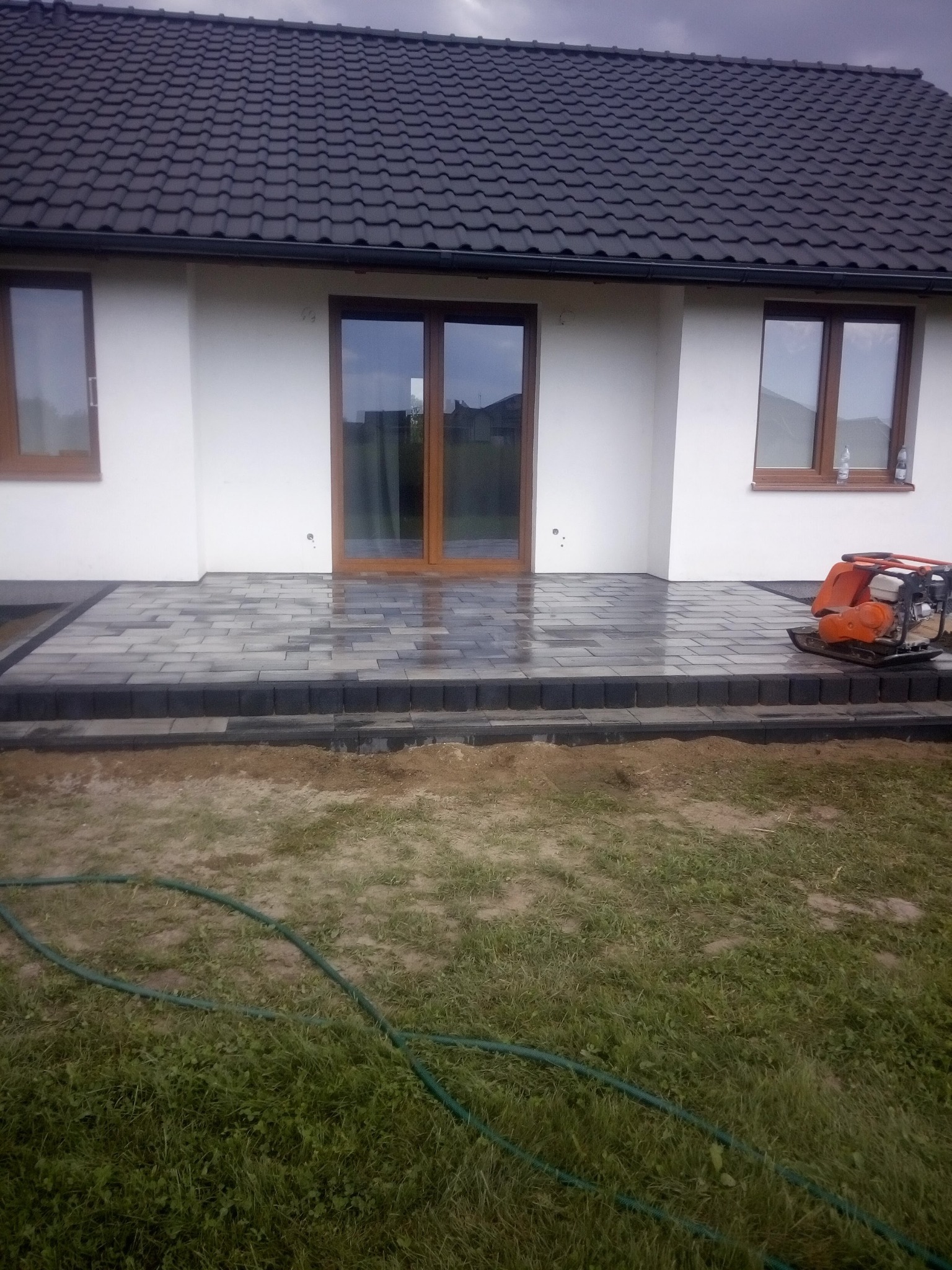 Nowo ułożony taras z szarej kostki brukowej przed domem z białymi ścianami i brązowymi oknami, z ubijarką wibracyjną obok, w tle pochmurne niebo.