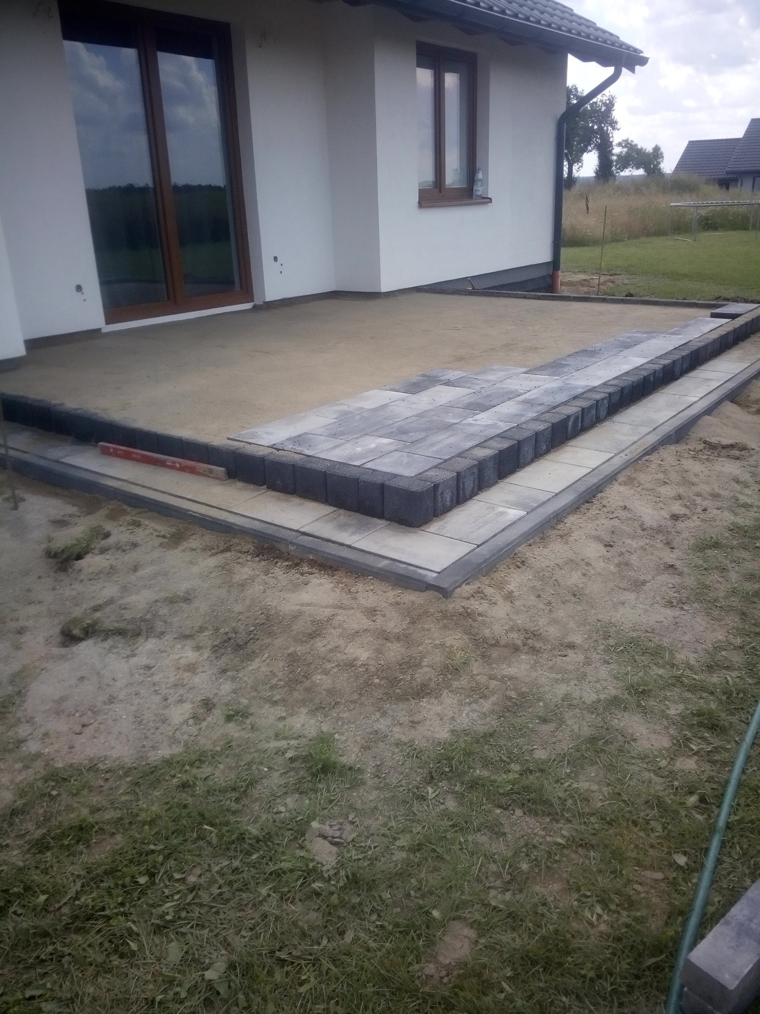 Przygotowanie podłoża i układanie obrzeży tarasu z kostki betonowej w kolorze szarym, widoczna poziomica, dom jednorodzinny w tle.