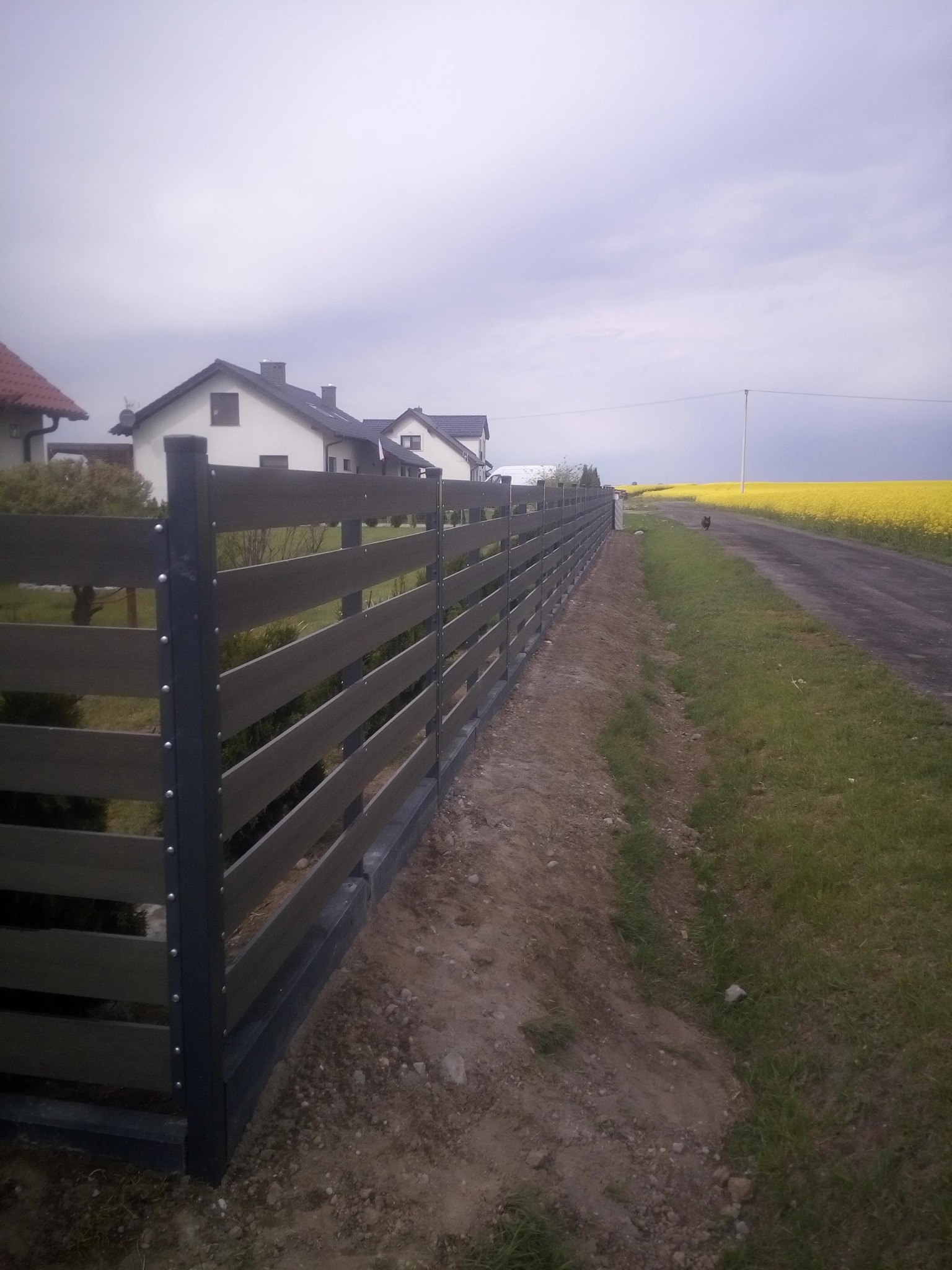 Długa, pozioma, drewniana, szara ogrodzenie panelowe z metalowymi słupkami, oddzielające posesję od polnej drogi z kwitnącym na żółto rzepakiem w tle.