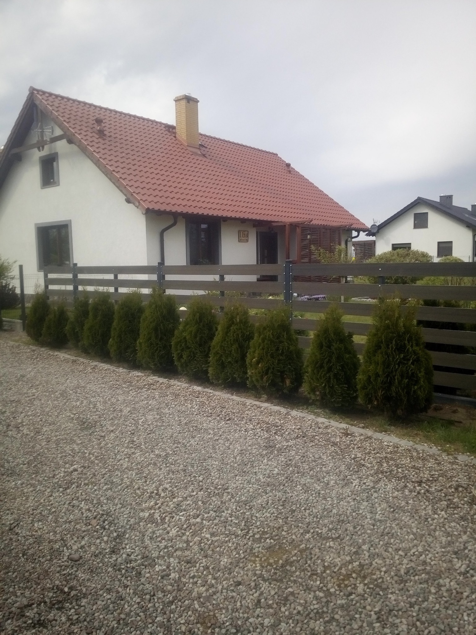 Nowoczesny dom jednorodzinny z brązowym dachem i drewnianym ogrodzeniem, ozdobiony rzędem małych, zadbanych tuj, wyłożony szarym żwirem.
