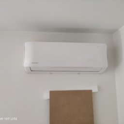 HYDROINSTAL-OZE Sławomir Łętocha - Biała klimatyzacja Rotenso zamontowana na ścianie, widoczny fragment kartonu zabezpieczającego ścianę podczas montażu. Minimalistyczny design, jasne wnętrze.