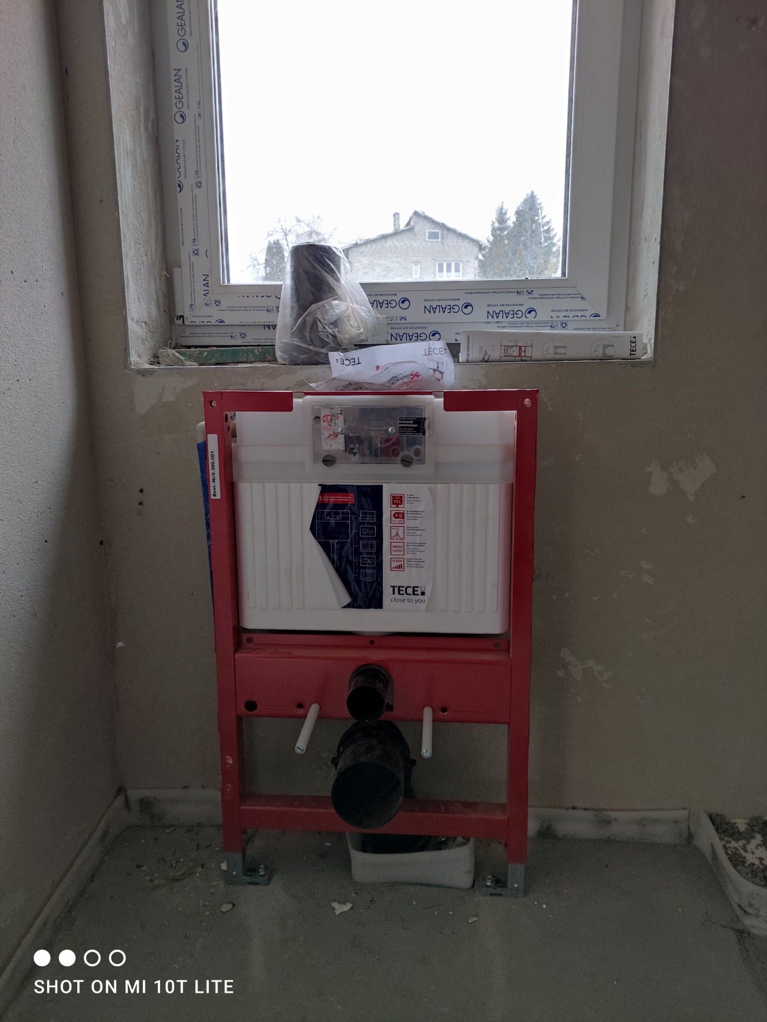 Czerwony stelaż podtynkowy TECE do WC zamontowany przy ścianie, widoczne przyłącza, na parapecie okna folia ochronna i instrukcja montażu.