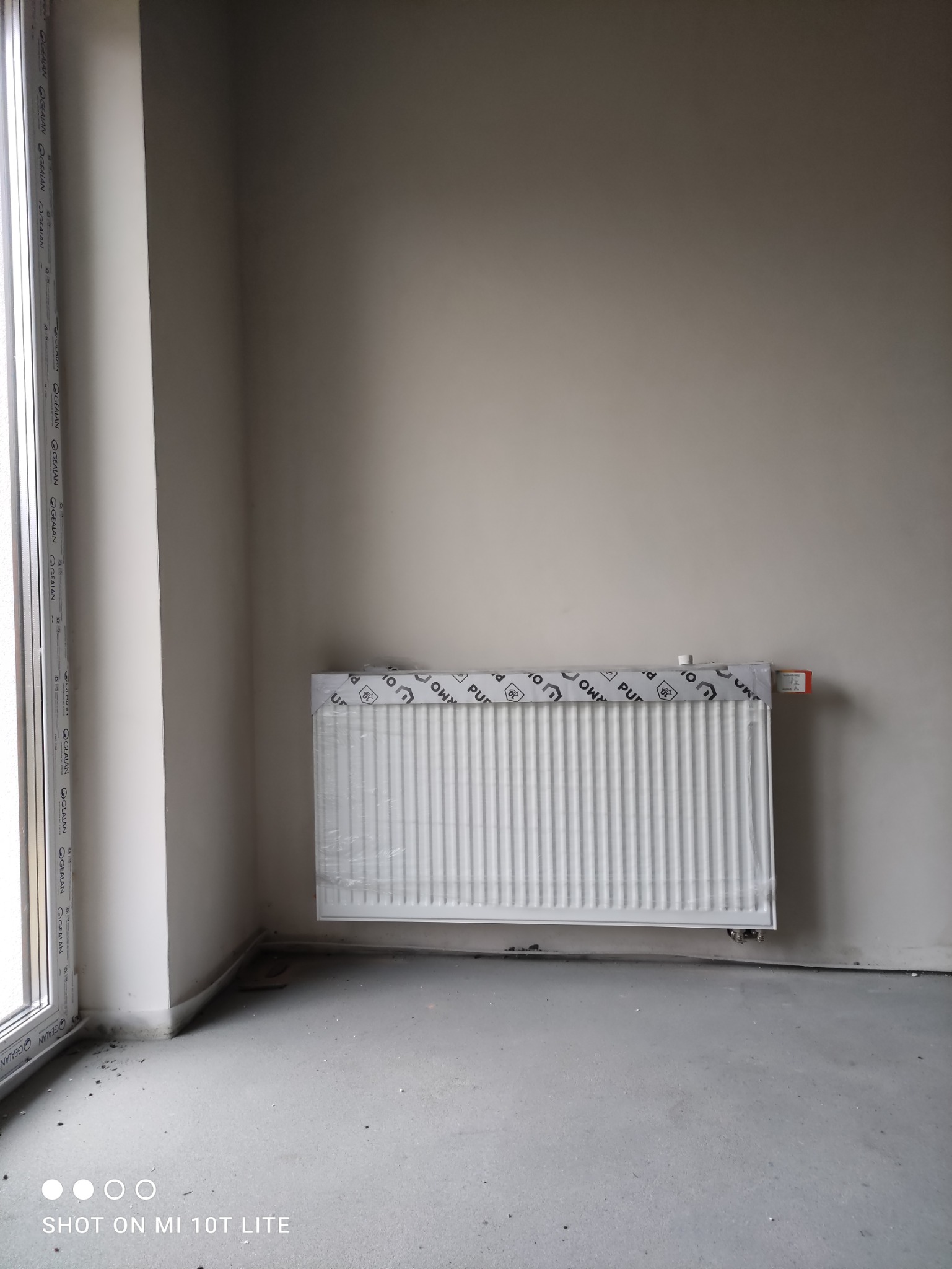 Nowy, biały grzejnik panelowy, jeszcze w folii ochronnej z nadrukiem, zamontowany na ścianie w surowym, szarym pomieszczeniu z oknem po lewej stronie. Widoczne przyłącza i fragment podłogi.