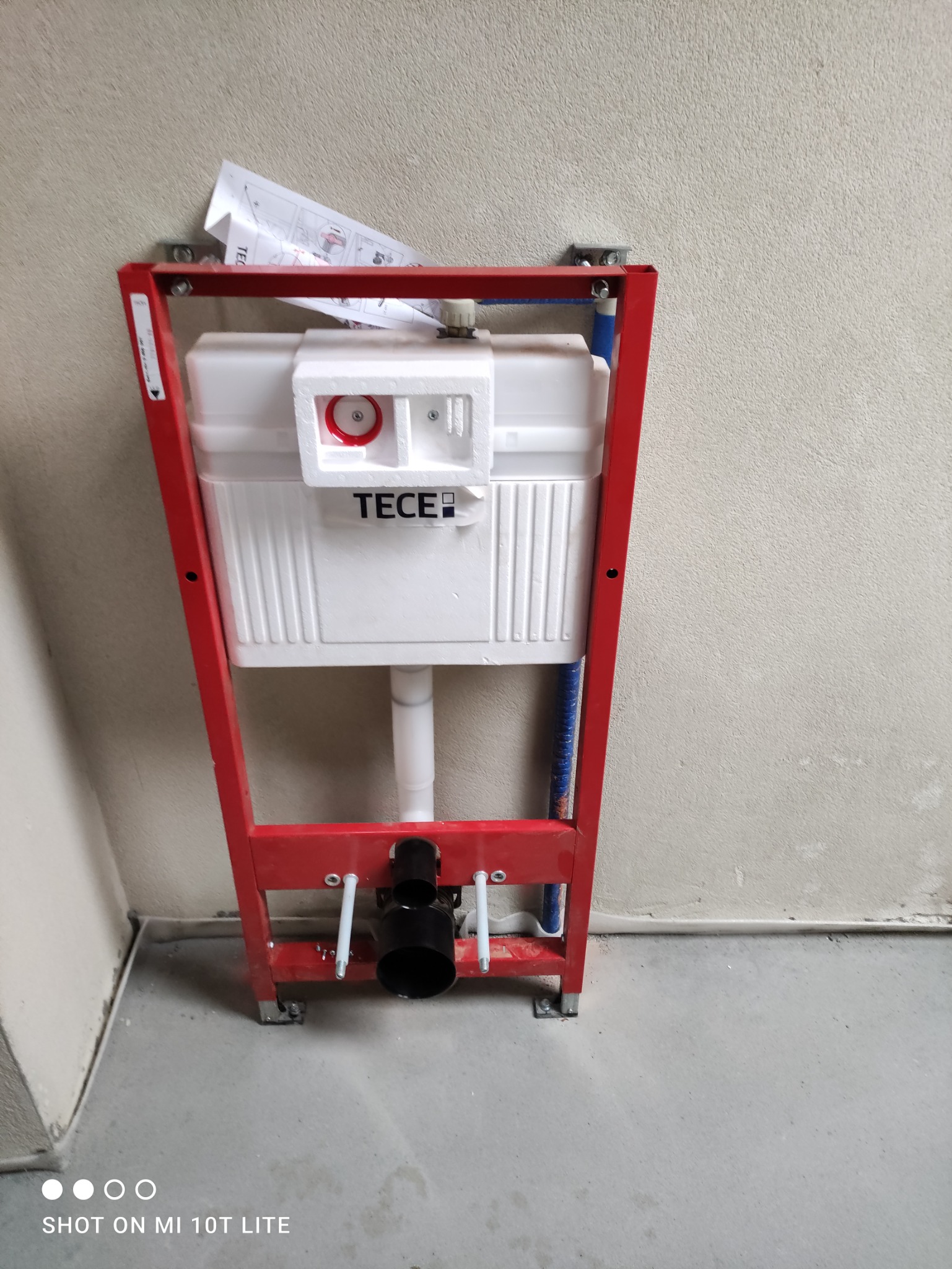 Stelaż podtynkowy WC marki TECE zamontowany w surowym stanie, widoczne niebieskie rury doprowadzające wodę oraz instrukcja montażu w tle.