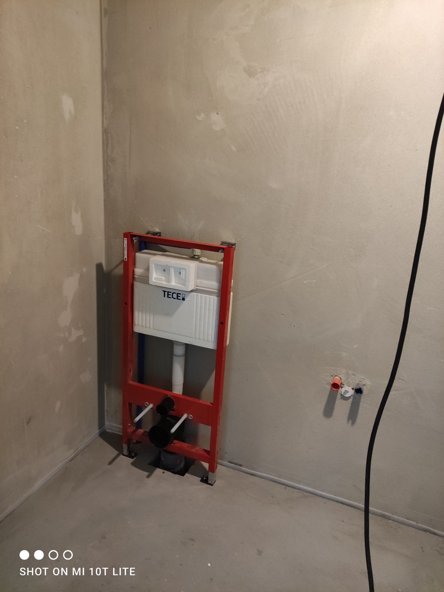 Stelaż podtynkowy TECE zamontowany w surowym pomieszczeniu, widoczne przyłącza wodne i kanalizacyjne w ścianie, ściany w stanie deweloperskim.