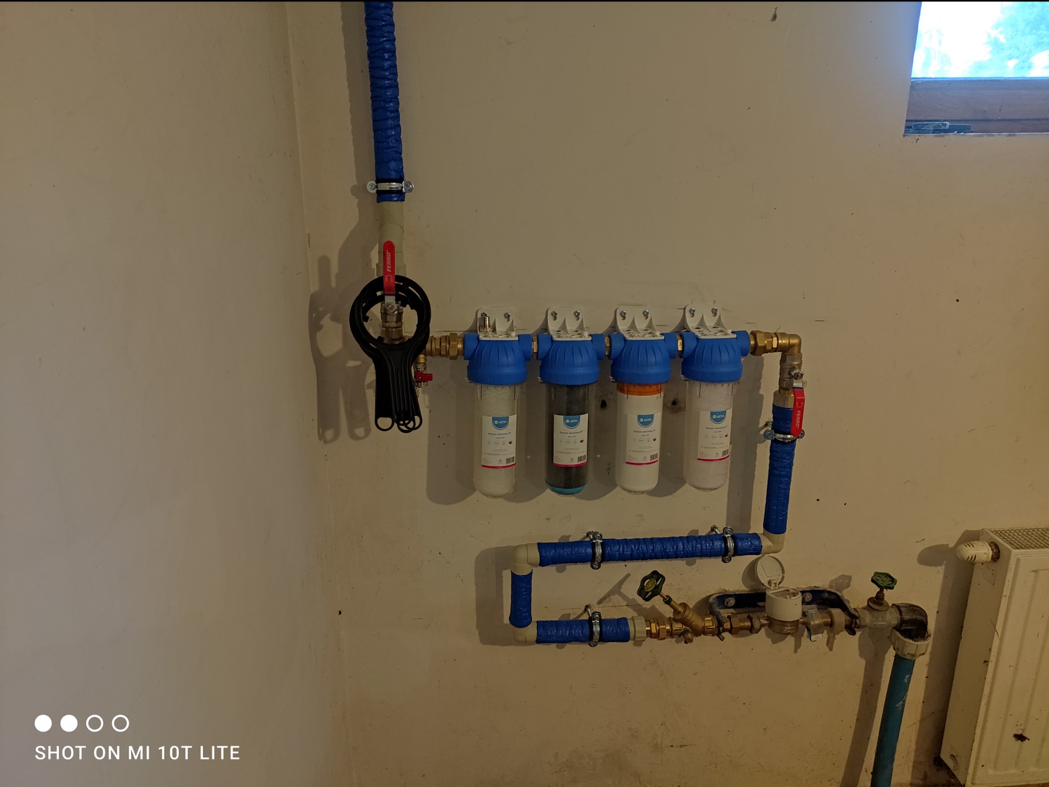 Cztery filtry wody z niebieskimi obudowami zamontowane na ścianie, połączone rurami w niebieskiej izolacji. Widoczny zawór i manometr. Ujęcie z dołu, w tle fragment okna.