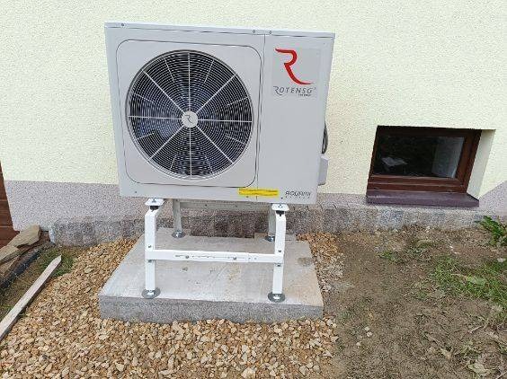 Zamontowana pompa ciepła Rotenso Aquami na betonowej podstawie z białym stelażem, widok z boku na tle ściany budynku z małym oknem piwnicznym.