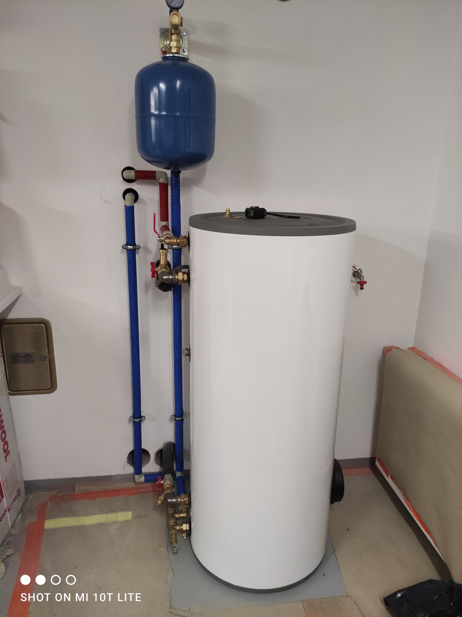 Bojler z niebieskim zbiornikiem wyrównawczym i rurami w izolacji, zamontowany w pomieszczeniu w Myślenicach. Widoczne przyłącza i armatura.