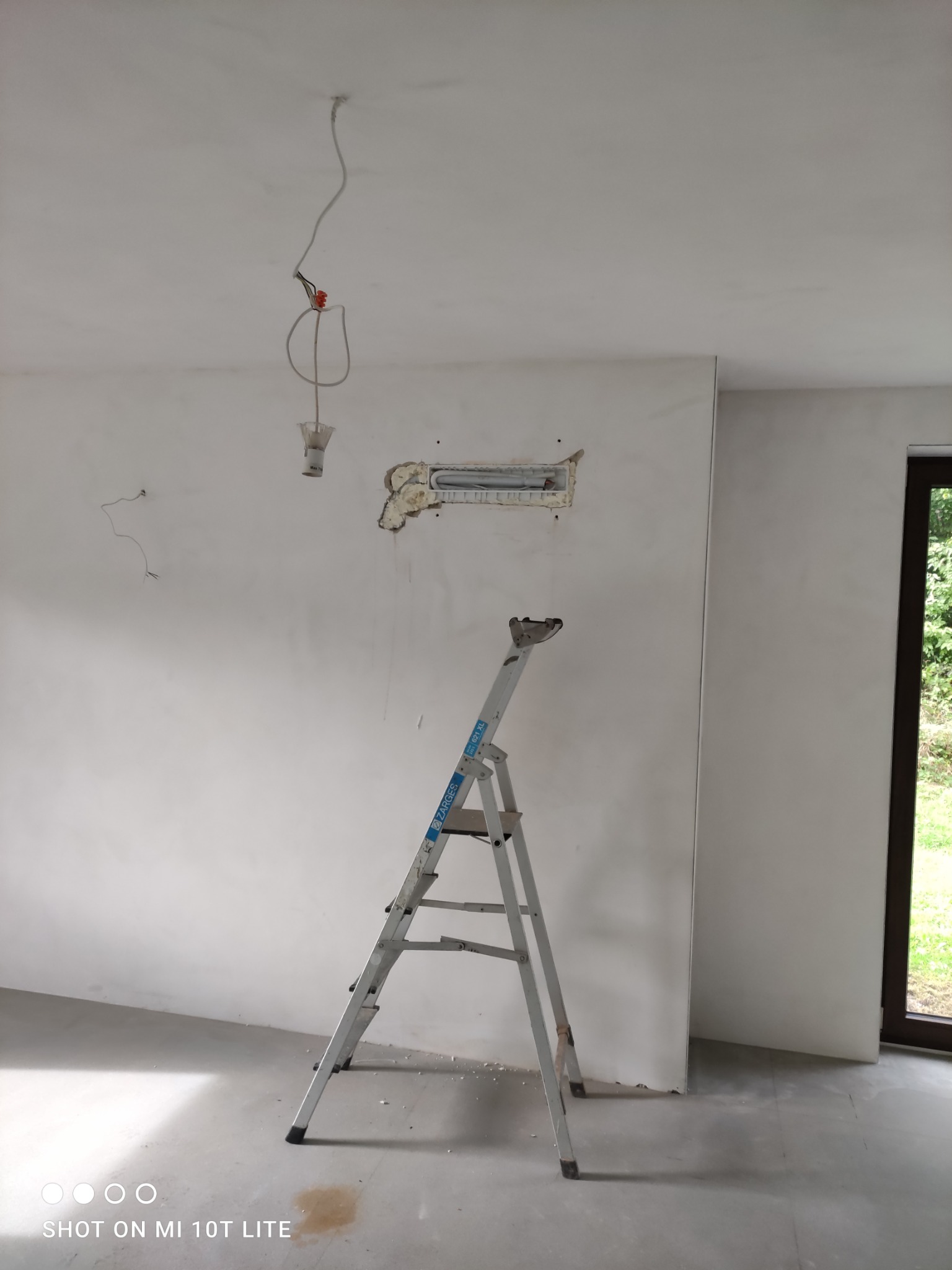 Ściana w trakcie remontu z wystającymi kablami i otworem na instalację, drabina oparta o ścianę, surowy stan wykończenia wnętrza, widok na zewnątrz przez okno.