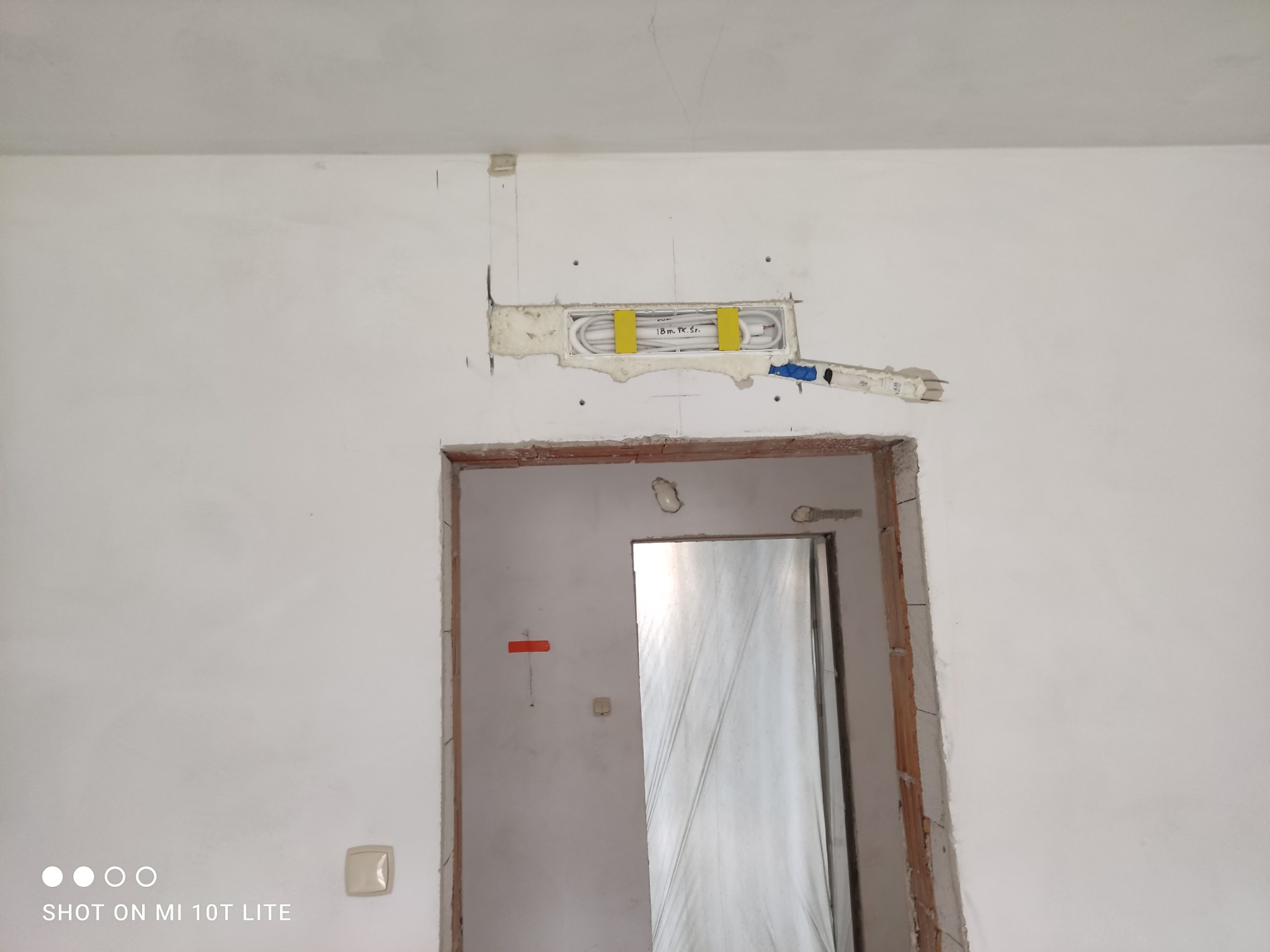 Wnęka w ścianie z ukrytą instalacją grzewczą, zabezpieczona żółtymi uchwytami, widoczna nad ościeżnicą drzwi w trakcie remontu. Białe ściany, surowy stan.