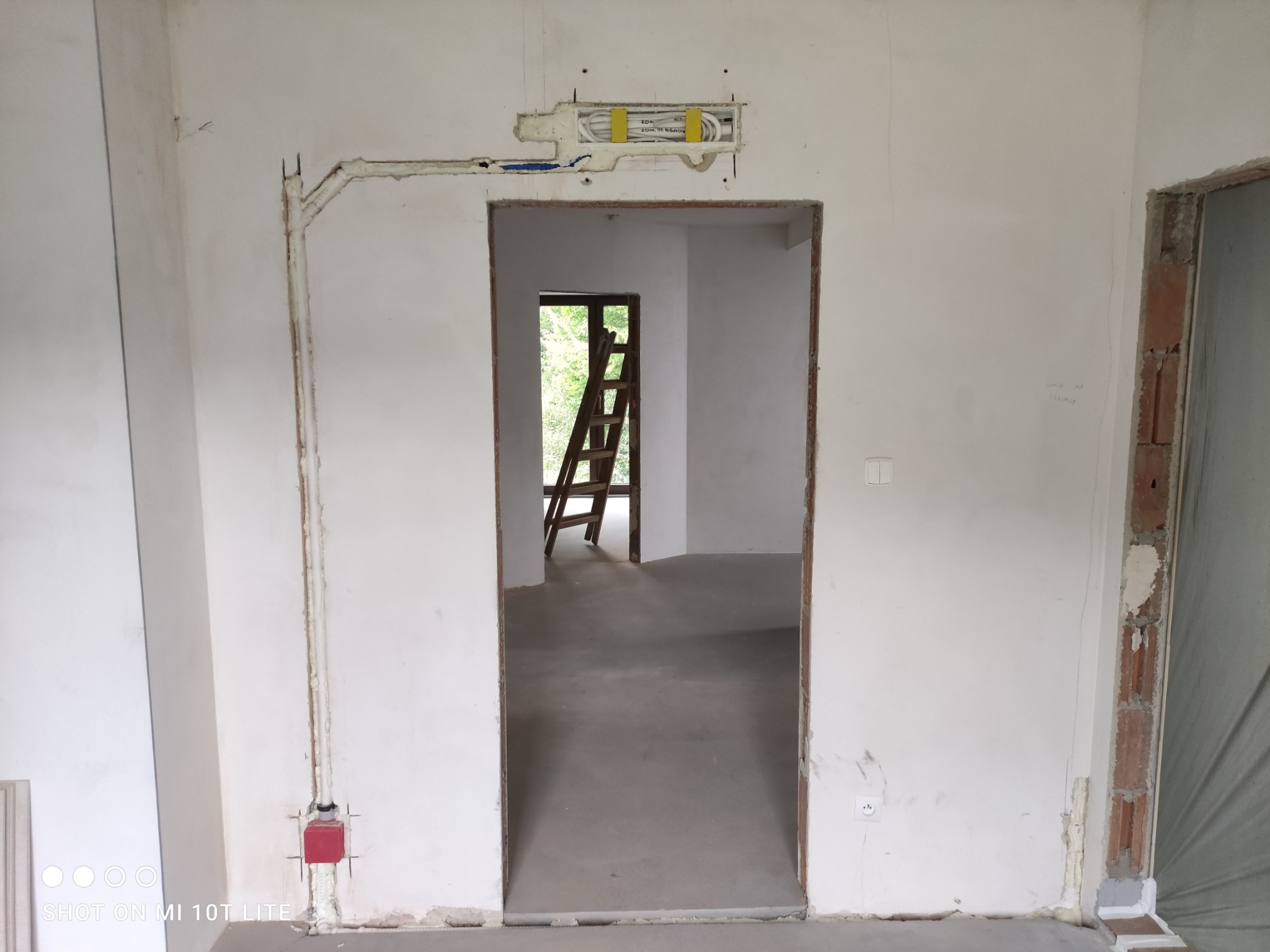 Instalacja elektryczna i hydrauliczna w surowym wnętrzu: rury przy ścianie, puszka elektryczna nad otworem drzwiowym, drabina w tle. Etap prac instalacyjnych.