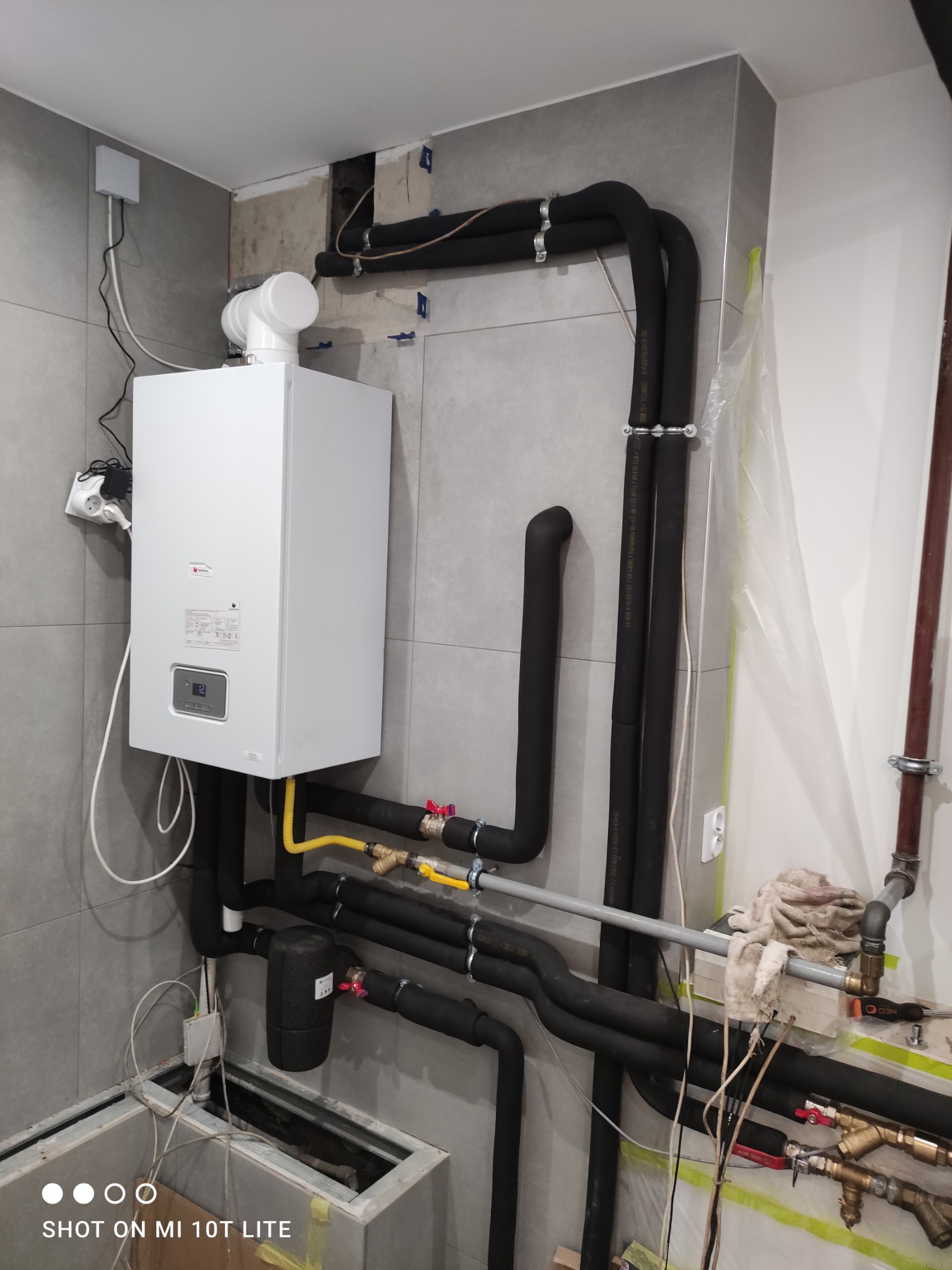 Biały piec gazowy na szarej ścianie z widocznymi rurami instalacji grzewczej, zaworami i elementami montażowymi. Widoczne narzędzia i elementy instalacji w tle.