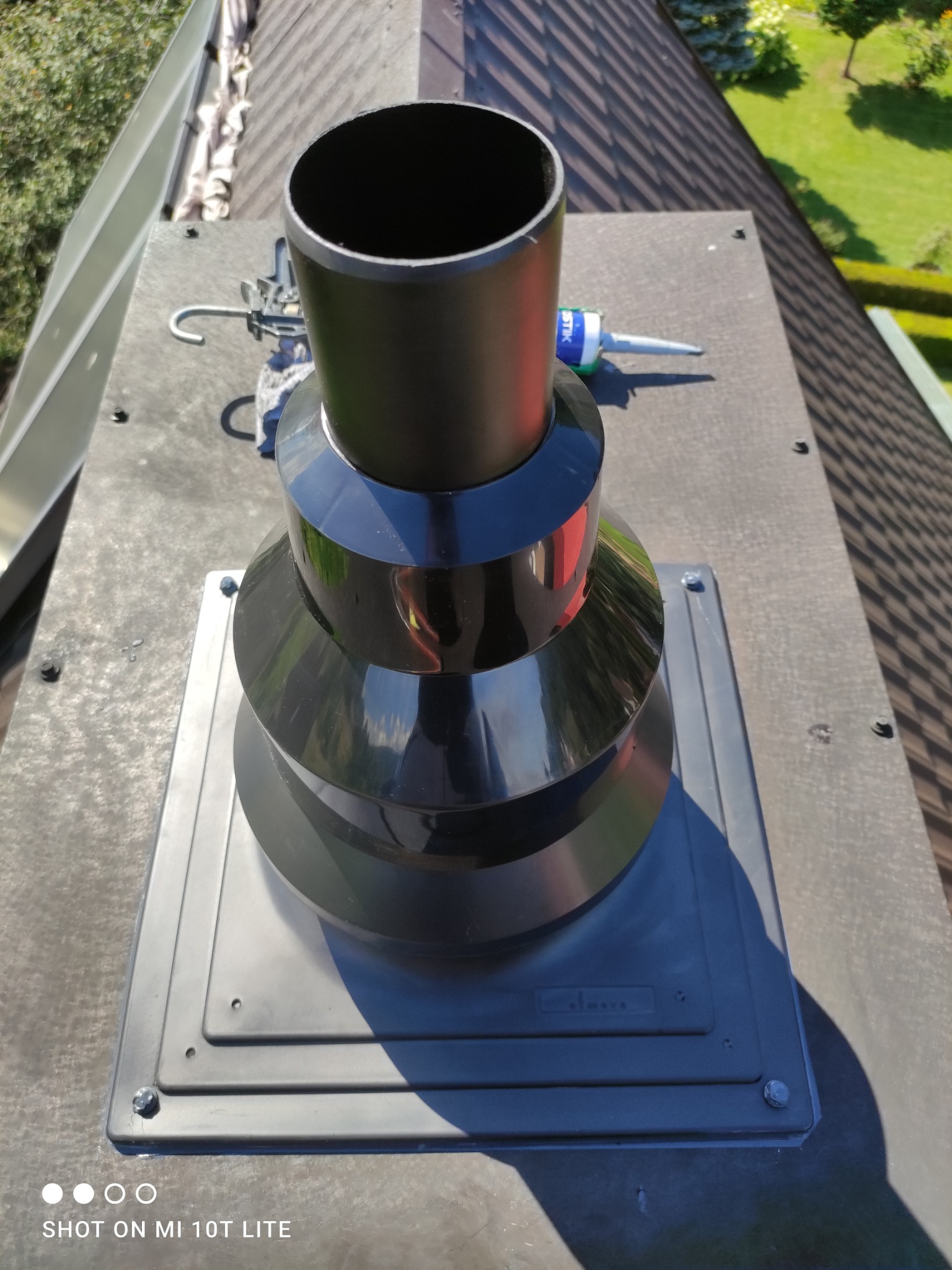 Czarny komin wentylacyjny na dachu z brązowej dachówki, widoczny silikon uszczelniający i narzędzia. Ujęcie z góry, w tle zieleń drzew.