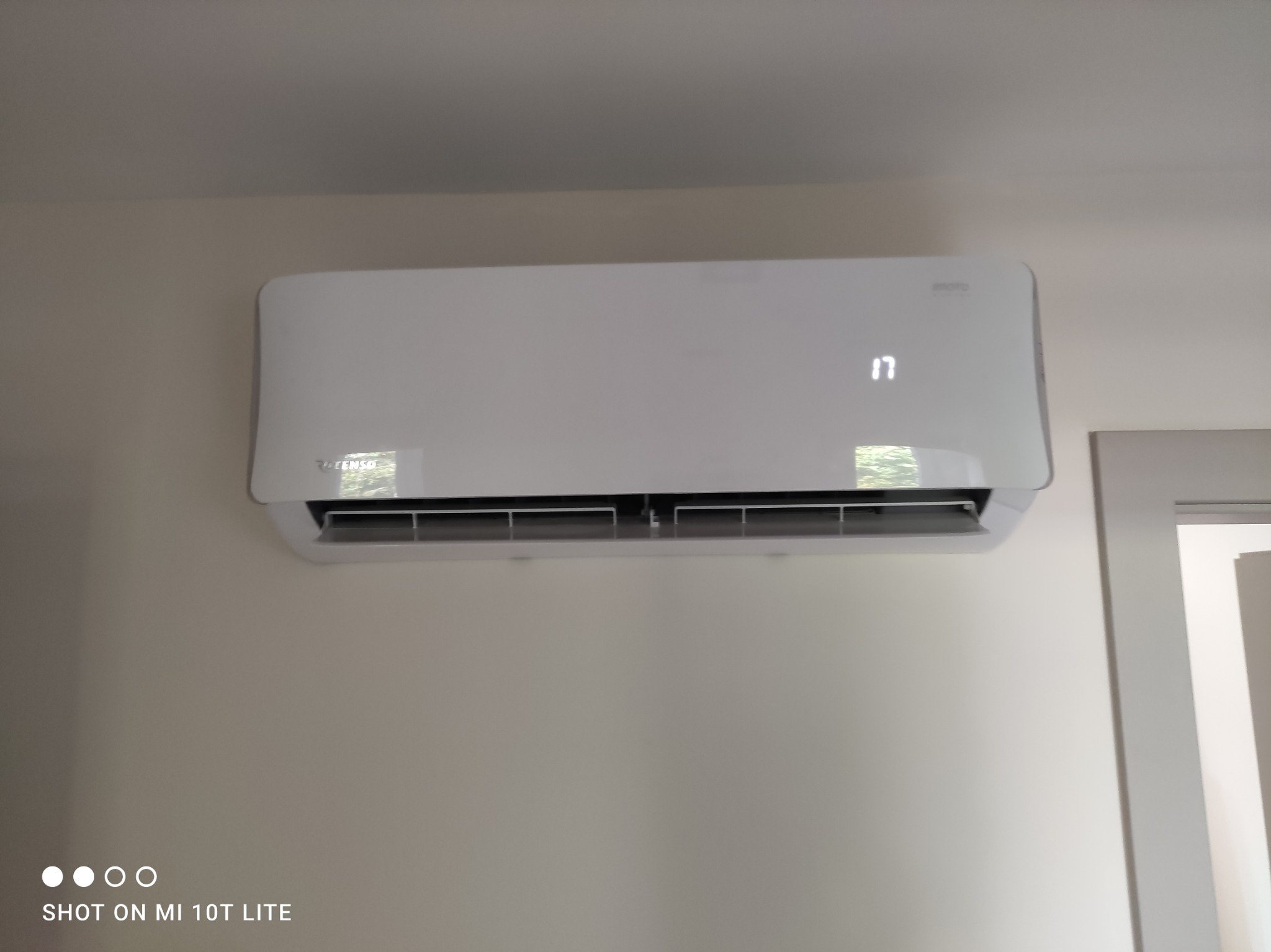 Biała klimatyzacja na ścianie, widoczny wyświetlacz z temperaturą 17 stopni, obok framuga drzwi. Minimalistyczny design, neutralne tło.