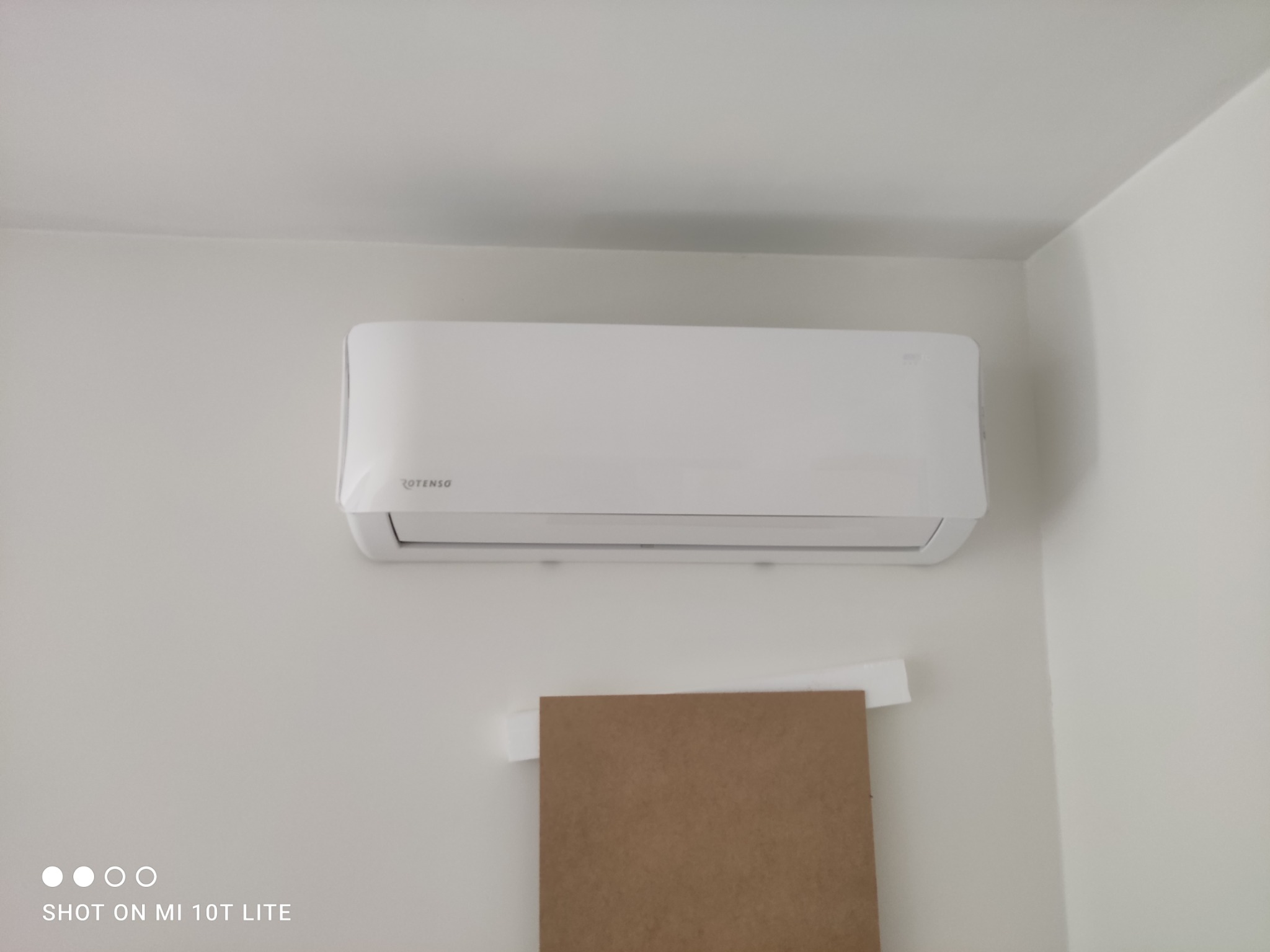 Biała klimatyzacja Rotenso zamontowana na ścianie, widoczny fragment kartonu zabezpieczającego ścianę podczas montażu. Minimalistyczny design, jasne wnętrze.
