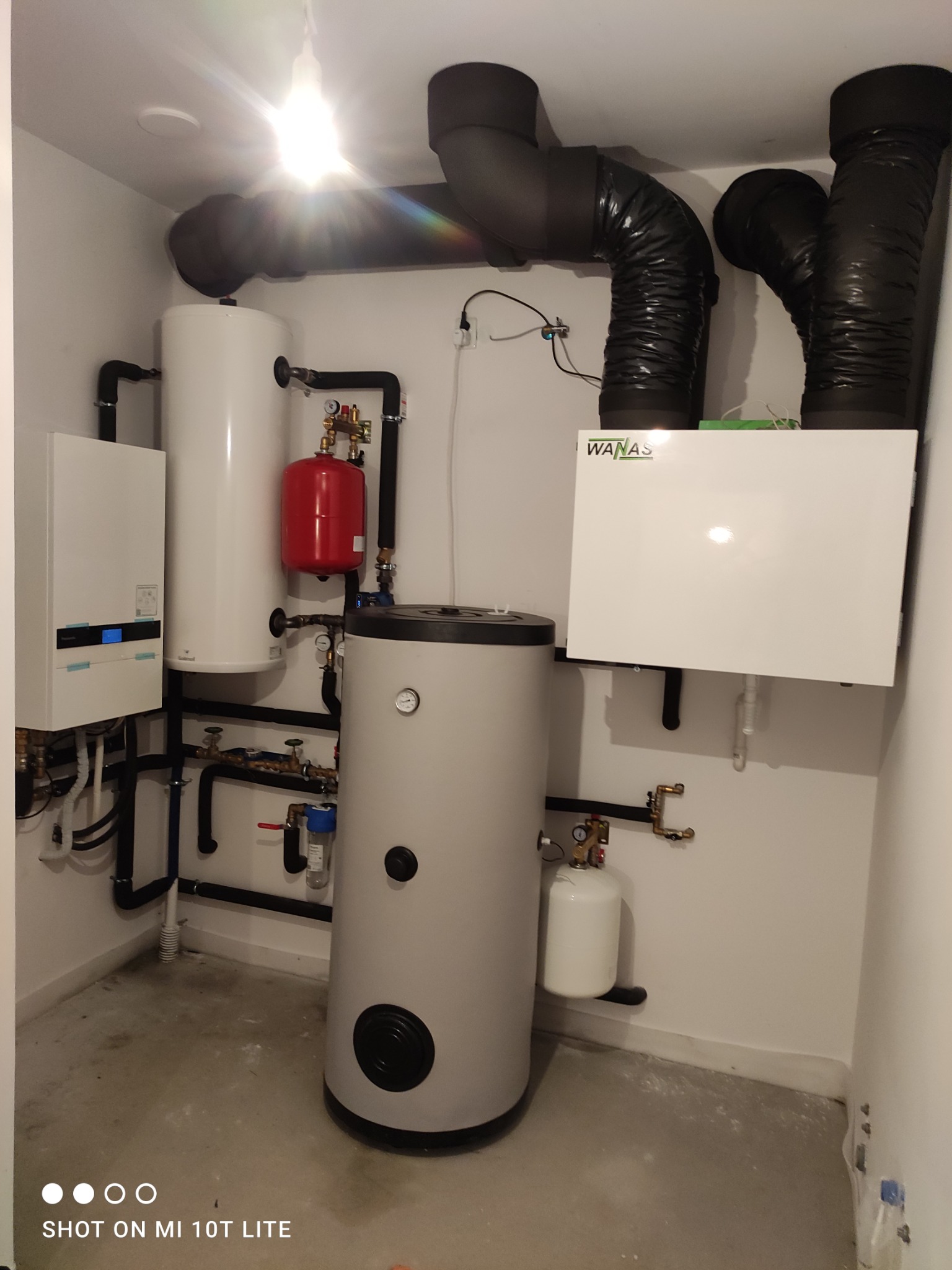Zestaw urządzeń grzewczych WANAS w kotłowni: biały zasobnik, czerwony zbiornik, szary bojler i biała pompa ciepła. Czarna instalacja rurowa. Oświetlenie sufitowe.