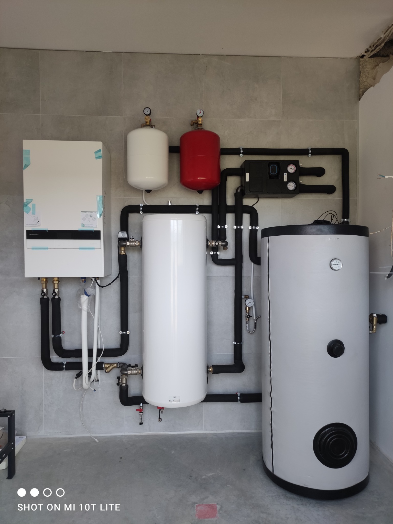 Zestaw urządzeń grzewczych: pompa ciepła, zbiorniki buforowe (biały i czerwony) oraz zasobnik ciepłej wody, połączone czarnymi rurami. Szare płytki na ścianie i podłodze.