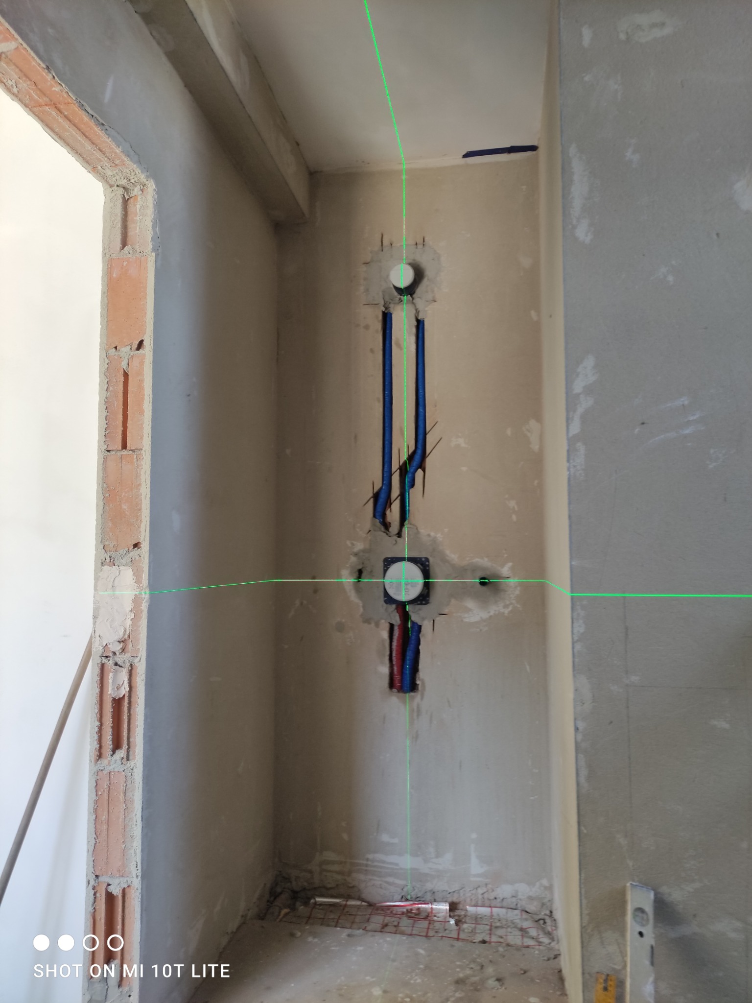 Ściana w trakcie prac hydraulicznych: widoczne niebieskie i czerwone rury, osadzone w ścianie, wyznaczane laserem. Surowy stan wykończenia, cegła i beton.