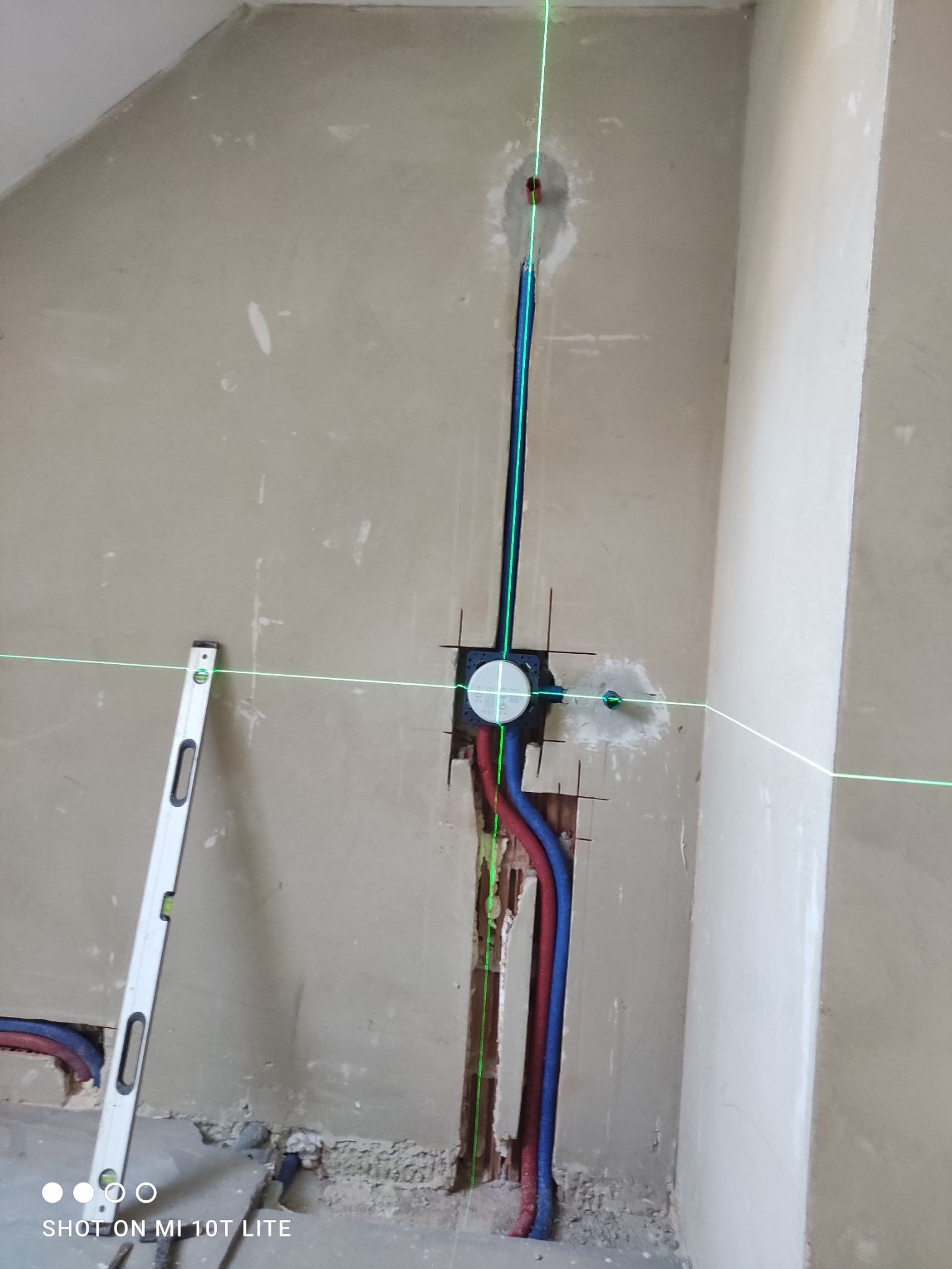 Ściana z instalacją hydrauliczną w trakcie montażu, widoczne rury w kolorze niebieskim i czerwonym, poziomica laserowa wyznacza pion i poziom.