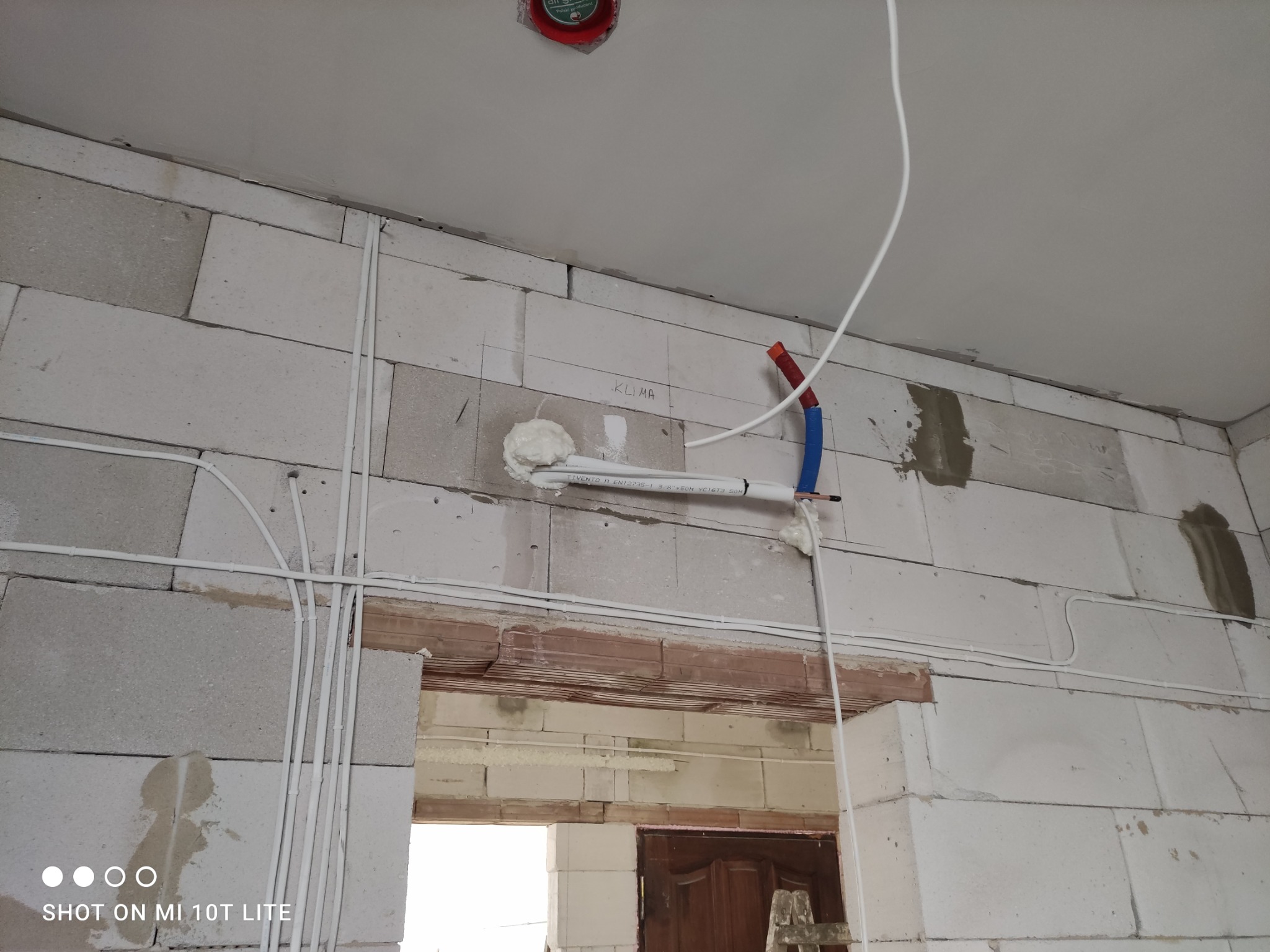 Instalacja rur klimatyzacji w surowym wnętrzu z białych bloczków. Widoczne przewody elektryczne i fragment otworu drzwiowego. Prace instalacyjne w trakcie realizacji.