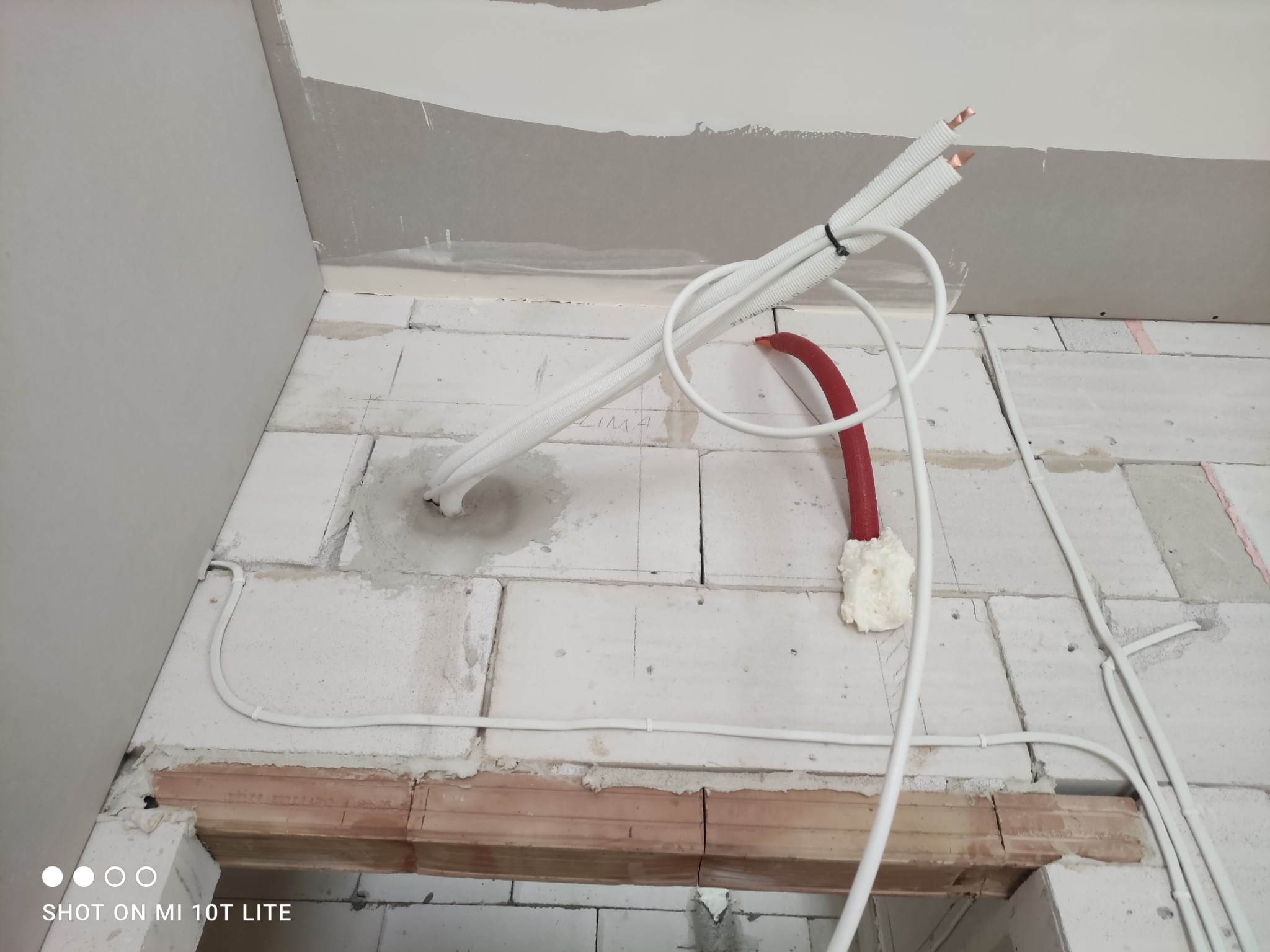 Białe i czerwone rury instalacyjne prowadzone wzdłuż ściany z betonu komórkowego, częściowo zakryte pianką montażową, widoczne przewody w białej osłonie.