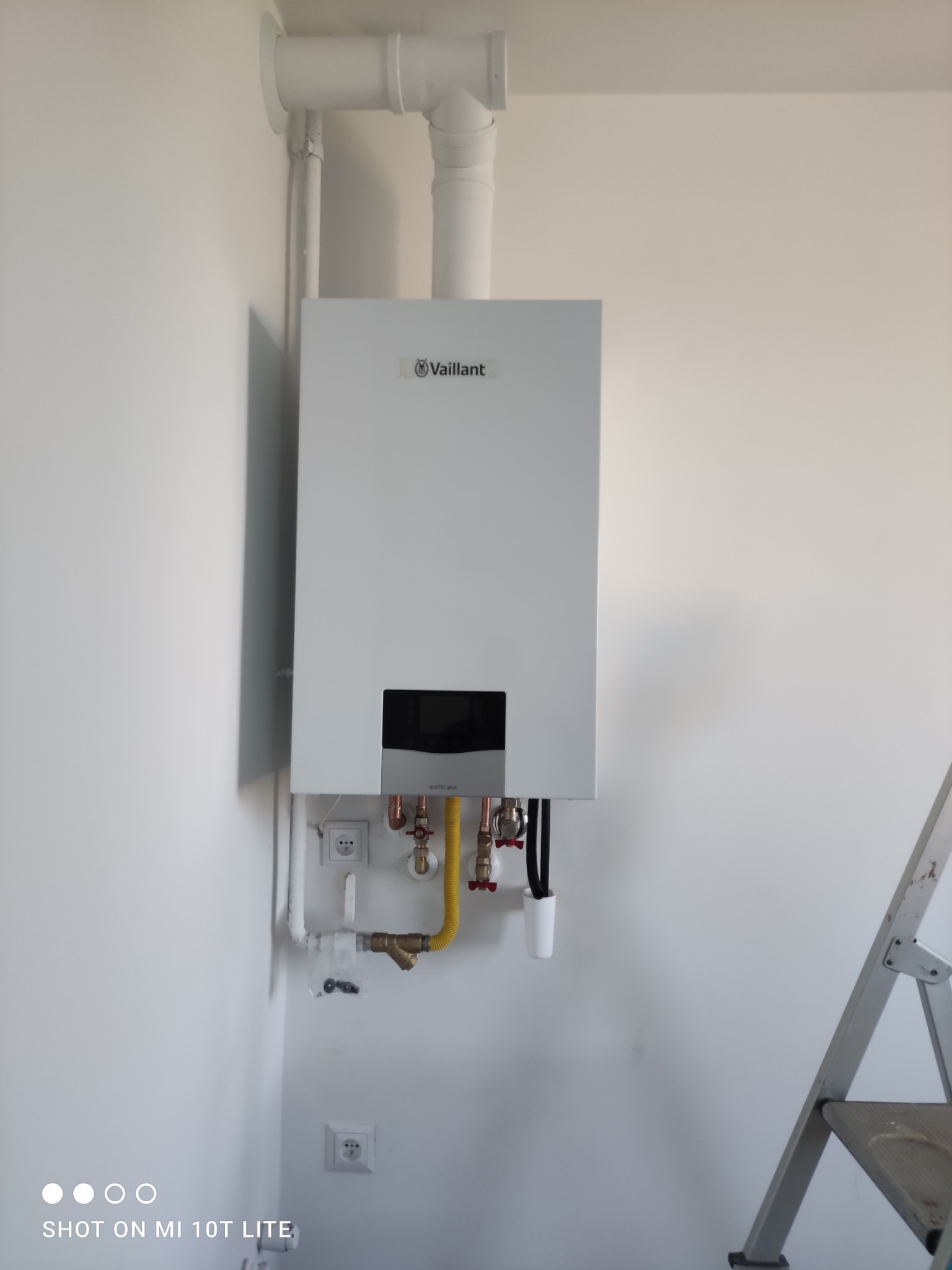 Biały, wiszący piec gazowy Vaillant na ścianie z widocznymi rurami i instalacją. Obok drabina. Ujęcie z dołu, podkreślające minimalistyczny design urządzenia.