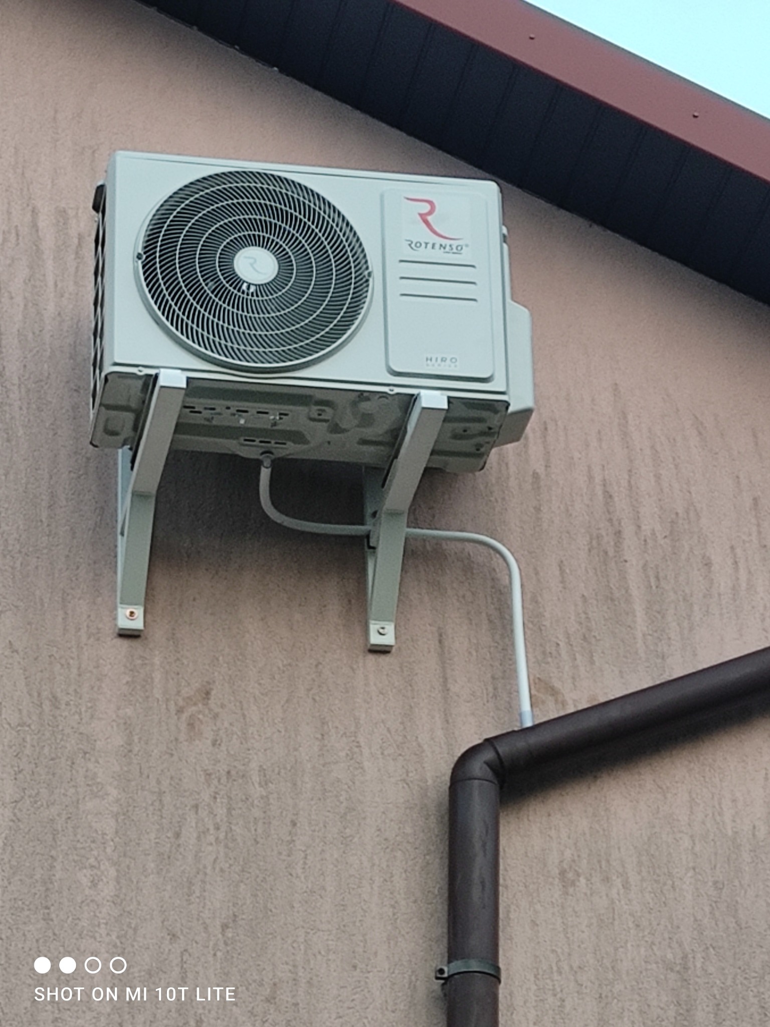 Zamontowana na elewacji zewnętrzna jednostka klimatyzacji Rotenso Hiro z widocznymi przewodami i rurą spustową. Ujęcie z dołu, lekko z prawej strony.