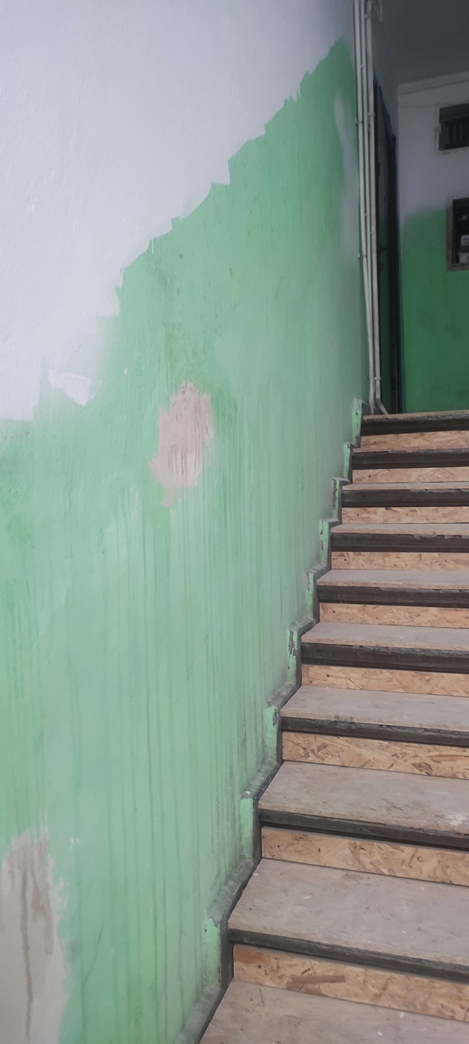 Ściana klatki schodowej w trakcie malowania na zielono, z widocznymi śladami po niedokładnym położeniu farby i fragmentami białej farby na górze, oraz surowe, drewniane schody z płyty OSB.