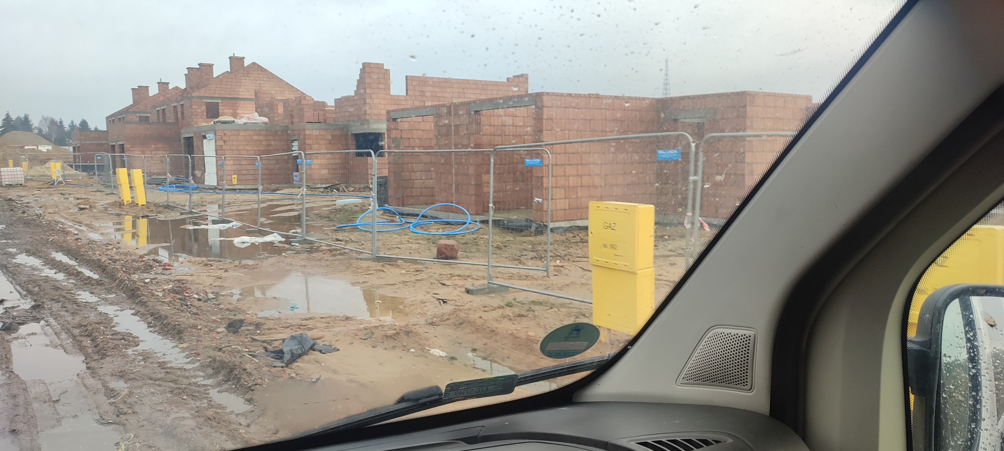 Surowy stan budowy domów z czerwonej cegły, widok z wnętrza pojazdu, z kałużami i błotem na placu budowy, zabezpieczone barierkami z niebieskimi rurami i żółtą skrzynką gazową.