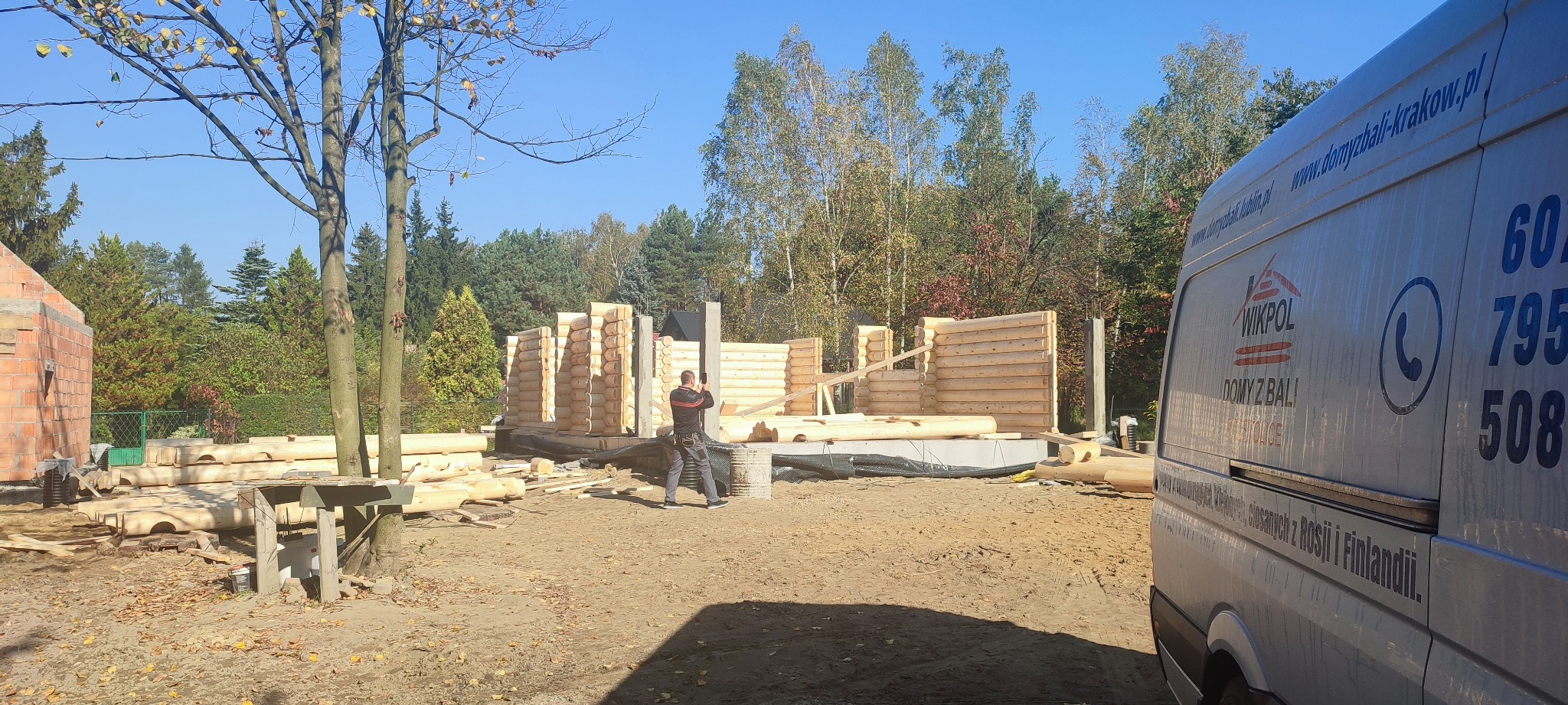 Wznoszenie ścian domu z bali, widoczny pracownik z telefonem, częściowo ukończona konstrukcja, fundament, teren budowy i samochód dostawczy firmy w tle.