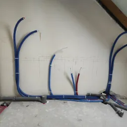Surowe wnętrze poddasza z widoczną instalacją hydrauliczną: niebieskie rury PEX prowadzone po ścianach i podłodze, szare rury kanalizacyjne z syfonem, widoczne przewody elektryczne, ściany...