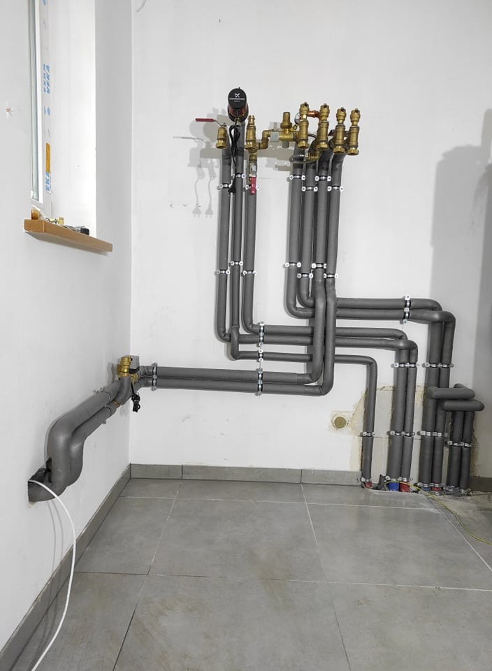 Szare, zaizolowane rury instalacji grzewczej z mosiężnymi zaworami i pompą obiegową, zamocowane do białej ściany na szarych płytkach podłogowych.