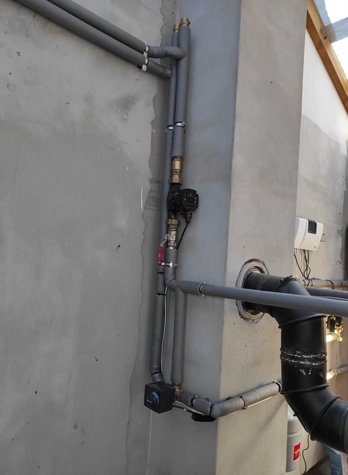 Szare, ocieplone rury instalacji grzewczej z zaworami i pompą obiegową zamontowane na surowej, szarej ścianie wewnętrznej budynku.