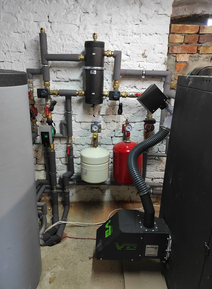 Instalacja kotłowni w Legnicy z piecem na ekogroszek, zbiornikami wyrównawczymi i izolowanymi rurami na tle ściany z cegły i białej cegły.