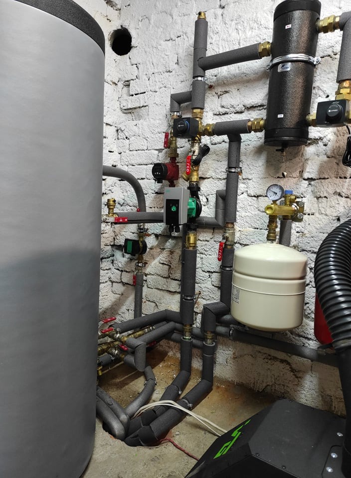 Szczegółowe ujęcie kotłowni z szarym zbiornikiem buforowym, rurami w izolacji termicznej, pompami obiegowymi, manometrem i białym naczyniem wzbiorczym na tle białej ściany z cegły.
