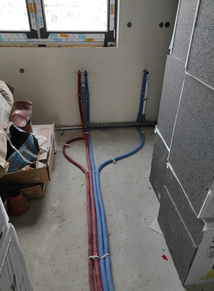 Wewnętrzna instalacja wodna z rurami w kolorze czerwonym i niebieskim, poprowadzona w surowym stanie budynku, widoczne elementy mocujące i fragment okna z profilem VEKA.