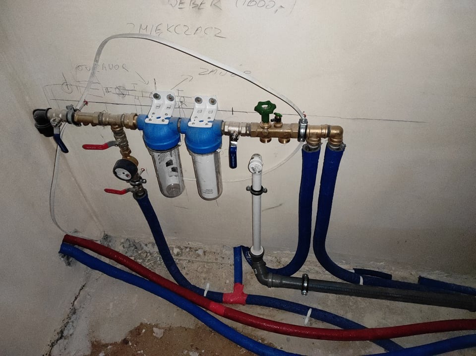 Zamontowany system filtrów wody z manometrem i rurami w izolacji termicznej, widoczne przyłącza i zawory, ściana z odręcznymi notatkami technicznymi.