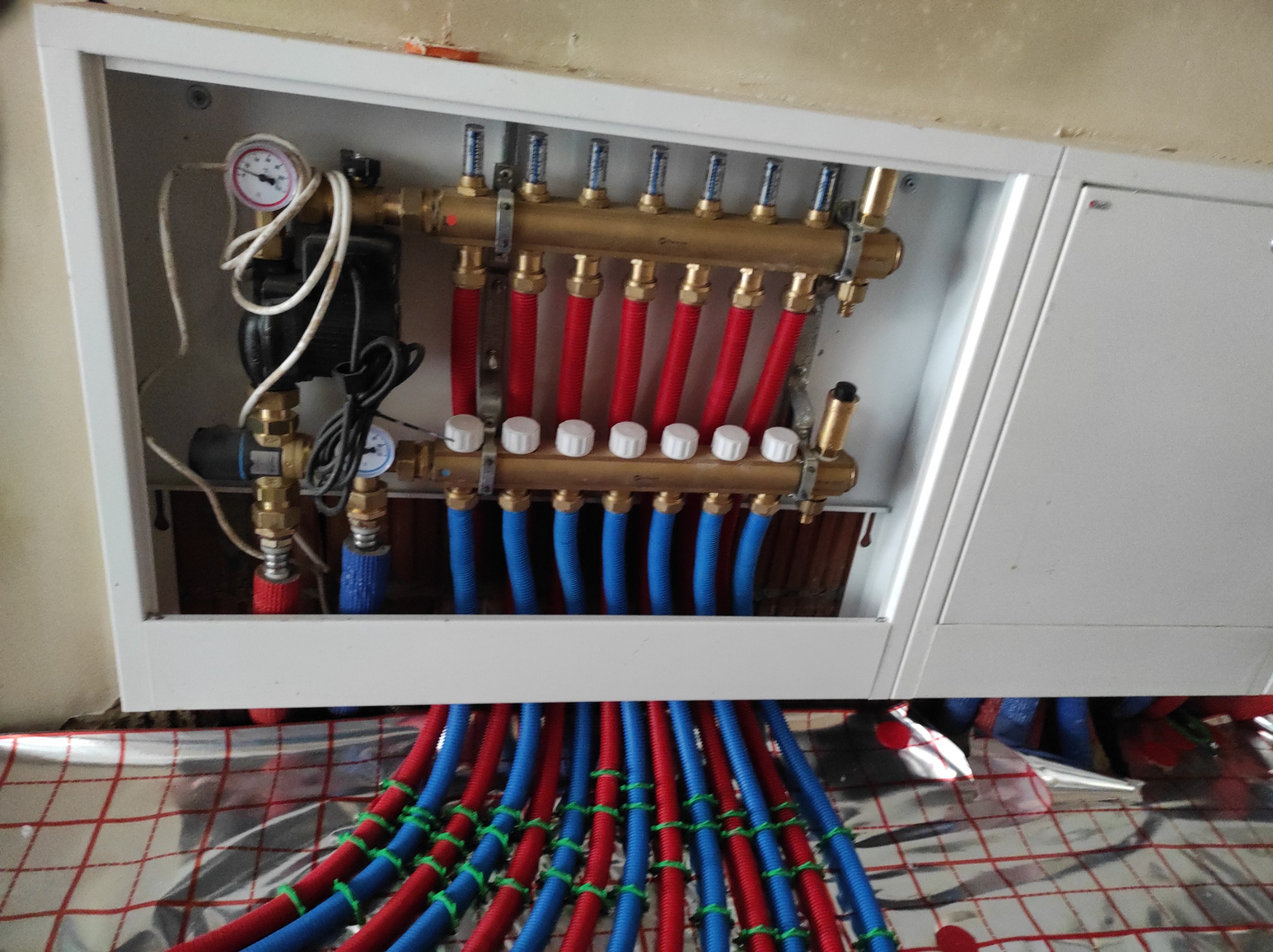 Rozdzielacz ogrzewania podłogowego w białej szafce, widoczne czerwone i niebieskie rury na folii izolacyjnej z siatką, manometr i zawory.