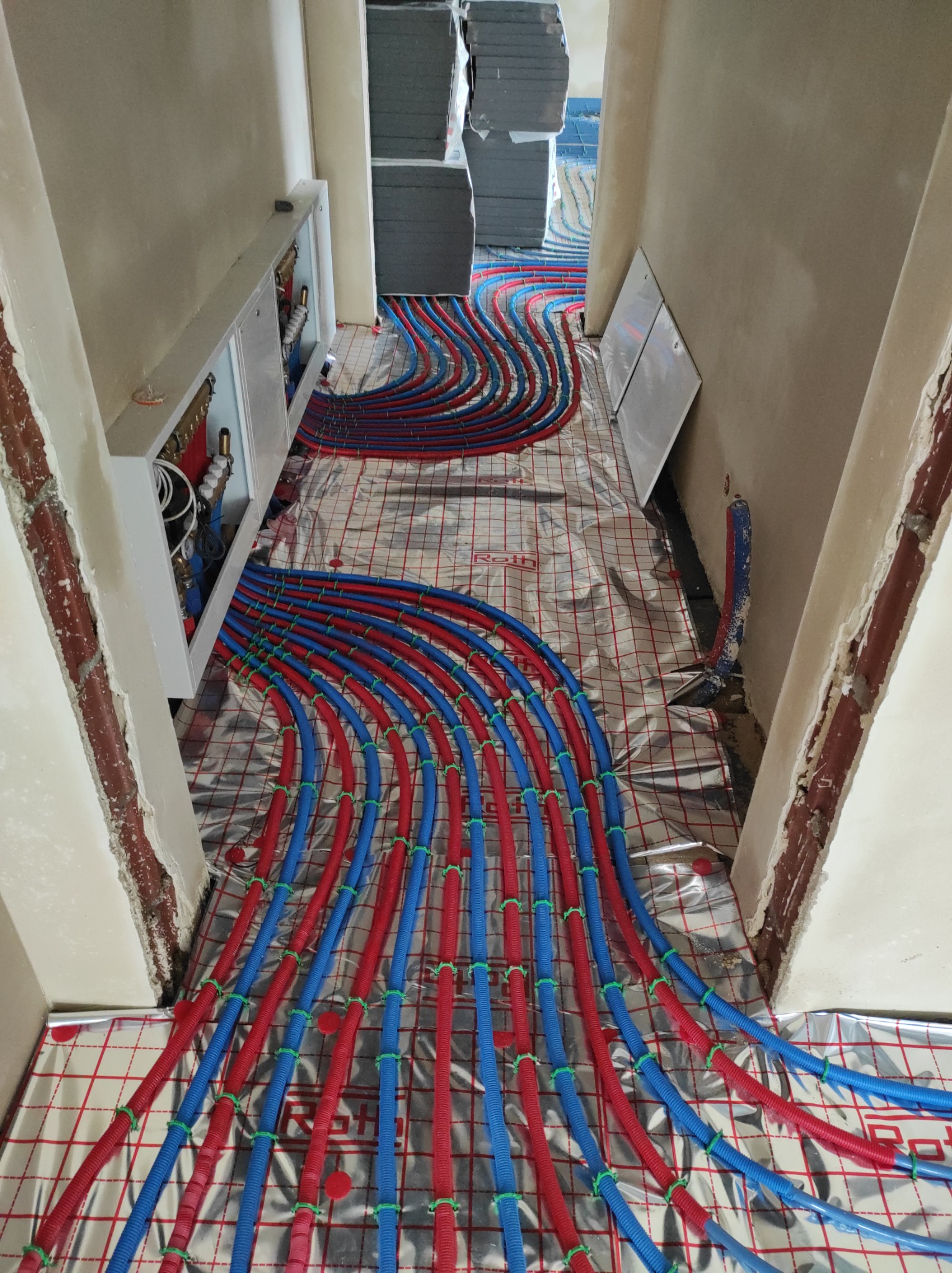 Rozłożone rury ogrzewania podłogowego w kolorze czerwonym i niebieskim, przymocowane zielonymi uchwytami do folii izolacyjnej z nadrukowaną siatką, widoczna szafka rozdzielcza na ścianie w stanie...