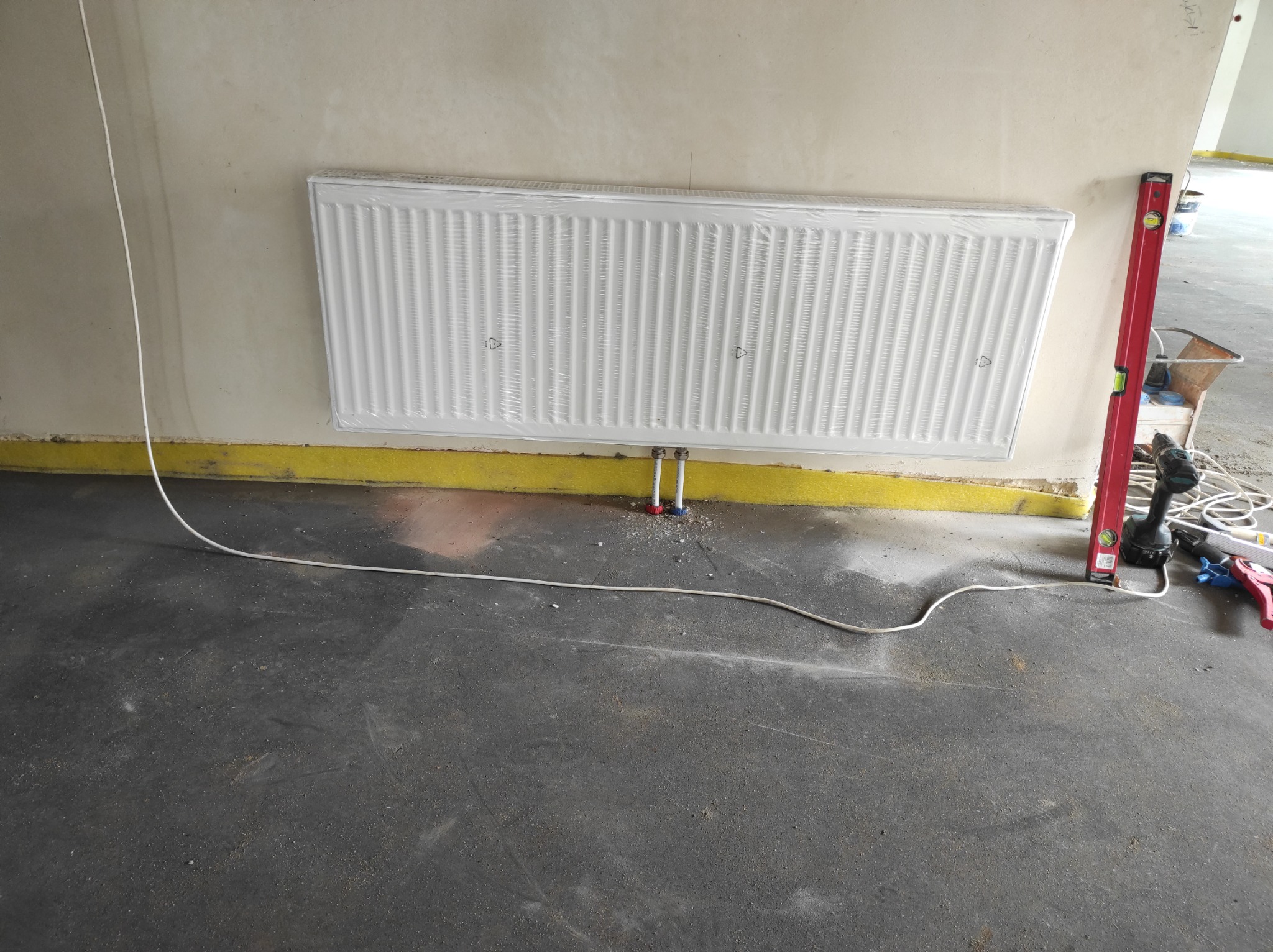 Nowy, biały grzejnik panelowy, jeszcze w folii ochronnej, zamontowany na ścianie. Widoczne rurki doprowadzające wodę, poziomica, wiertarka i inne narzędzia na podłodze. Surowy stan wykończenia...