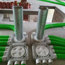 Dwa rozdzielacze wentylacyjne z podłączonymi zielonymi przewodami karbowanymi i pionowymi metalowymi rurami z oznaczeniami 'N' i 'W', w surowym wnętrzu budynku.