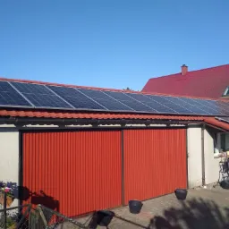 Panele słoneczne na dachu garażu z czerwoną bramą, w tle dom z czerwoną dachówką i błękitne niebo.