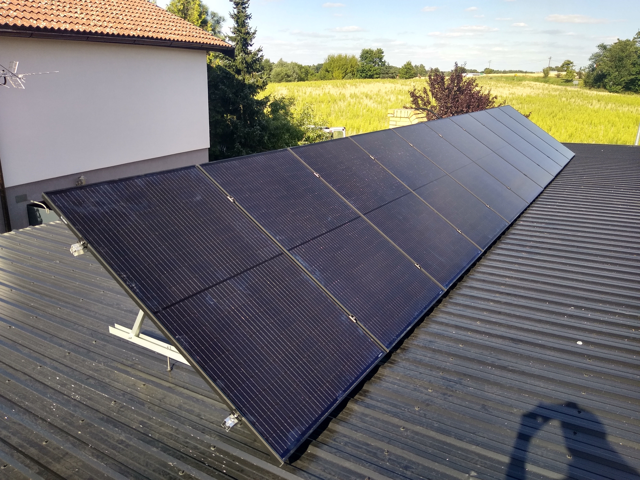 Rząd czarnych paneli słonecznych na metalowym dachu domu jednorodzinnego, widok na zielone pole w tle.