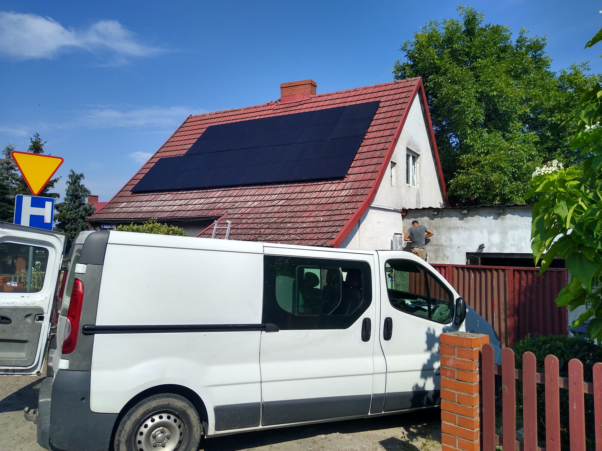 Montaż paneli na dachu domu jednorodzinnego z czerwonej dachówki, widoczny biały bus instalacyjny i pracownik na drabinie