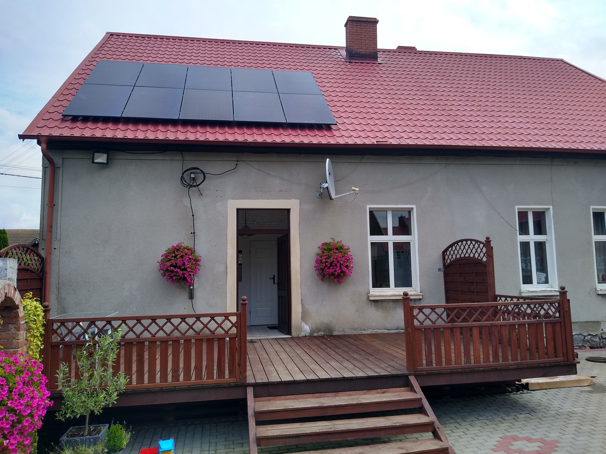 Dom jednorodzinny z czerwonym dachem i panelami słonecznymi, widoczne kwiaty w donicach na ścianie i drewniany taras z przodu.